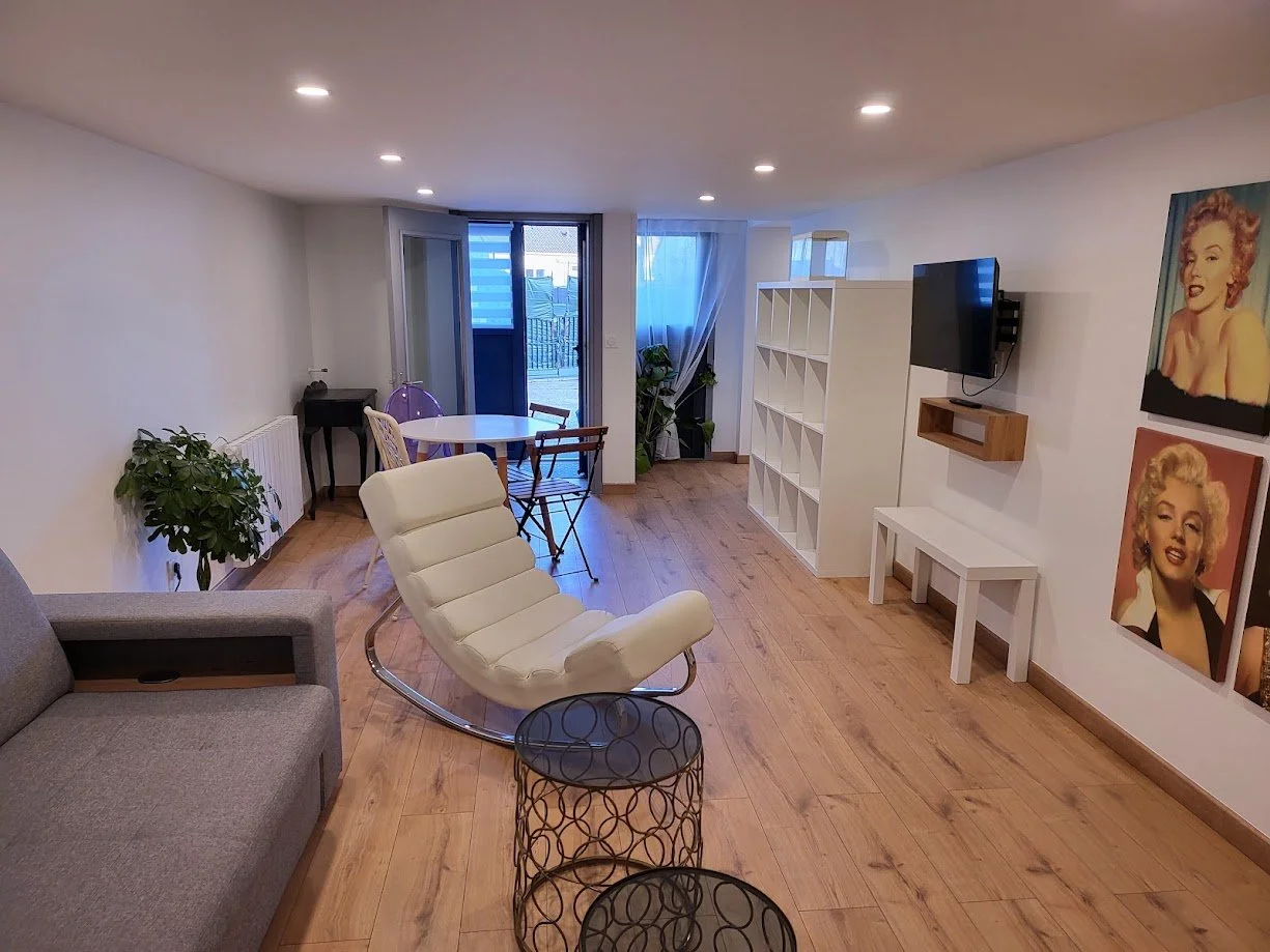 Salon avec un fauteuil blanc inclinable, un canapé gris, des tables, une étagère blanche, une télévision murale, des lampes encastrées au plafond, des posters de Marilyn Monroe sur le mur, une porte-fenêtre menant à un balcon, des plantes vertes, un 