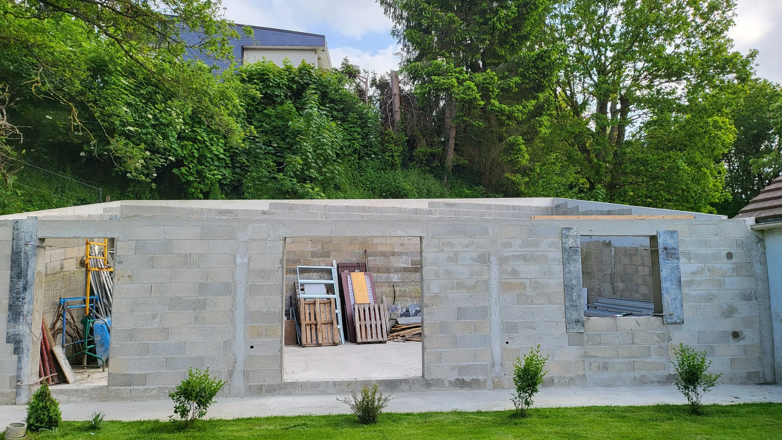 Construction d'un mur en briques avec deux fenêtres et un espace de porte, sur un terrain herbeux, avec des arbres verts en arrière-plan.