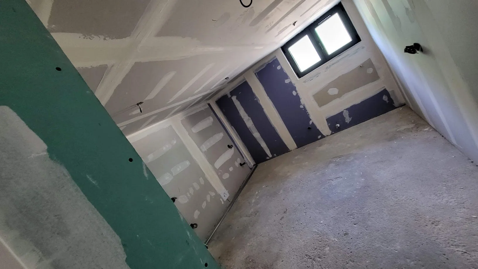 Une pièce en construction avec murs en plaques de plâtre non peintes et un sol en béton, une fenêtre au mur et quelques installations électriques en cours.