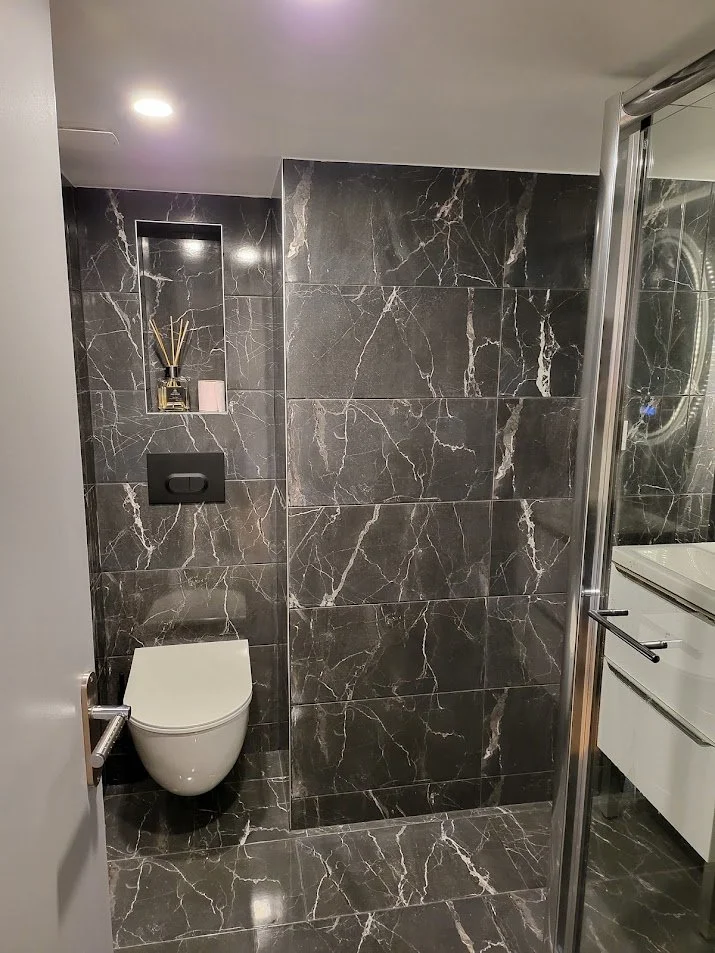 Salle de bain moderne avec murs et sol en carrelage noir à veins blanches, toilette suspendue blanche, étagère intégrée avec diffuseur de parfum et bougie, cabine de douche avec porte en verre, meuble lavabo blanc avec miroir.