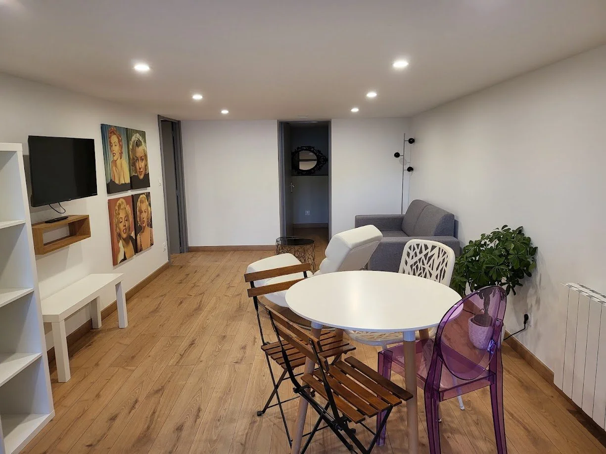 Salon avec une télévision murale, quatre portraits de Marilyn Monroe, un canapé gris, une table ronde blanche, une chaise en plastique violette, une chaise en bois, un fauteuil blanc, une plante verte, des murs blancs et un sol en bois.