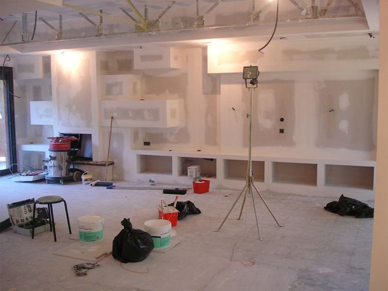 Une pièce en cours de rénovation avec des murs en plâtre, des meubles de chantier, et un éclairage de travail. Le sol est recouvert de poussière et de matériaux de construction.