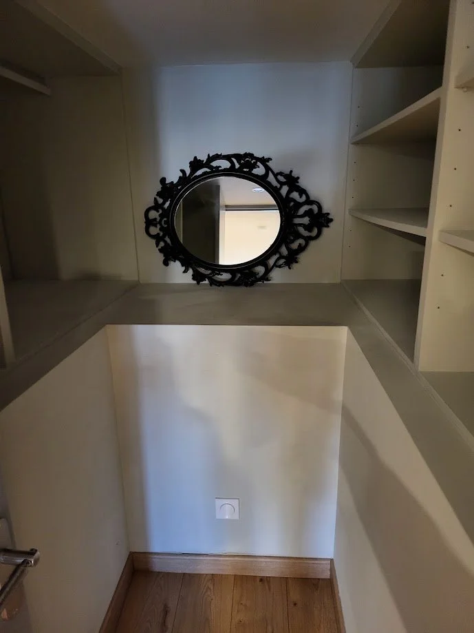 Un miroir noir au cadre décoratif est fixé au mur dans une pièce avec des étagères blanches. La pièce a un sol en bois et une prise électrique visible en bas.
