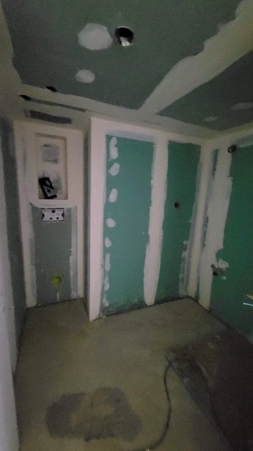 Pièce en rénovation avec murs et plafond en plaques de plâtre, pièces de coupe dans le plafond, et sol en béton, avec câbles électriques visibles.