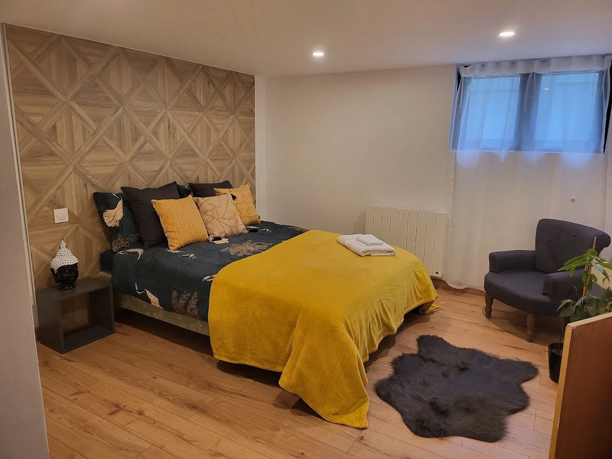 Chambre avec lit, coussins jaunes et noirs, couverture jaune, tapis en fourrure gris, fauteuil gris, fenêtre avec rideaux blancs, décoration en bois sur le mur derrière le lit, petite table avec décoration en haut, plante verte