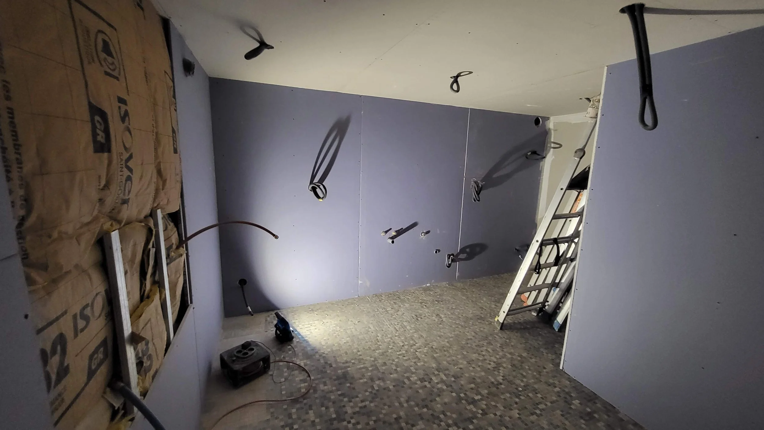 Salle en rénovation avec murs en placo, câbles électriques visibles et outillage sur le sol. Sol en mosaïque.