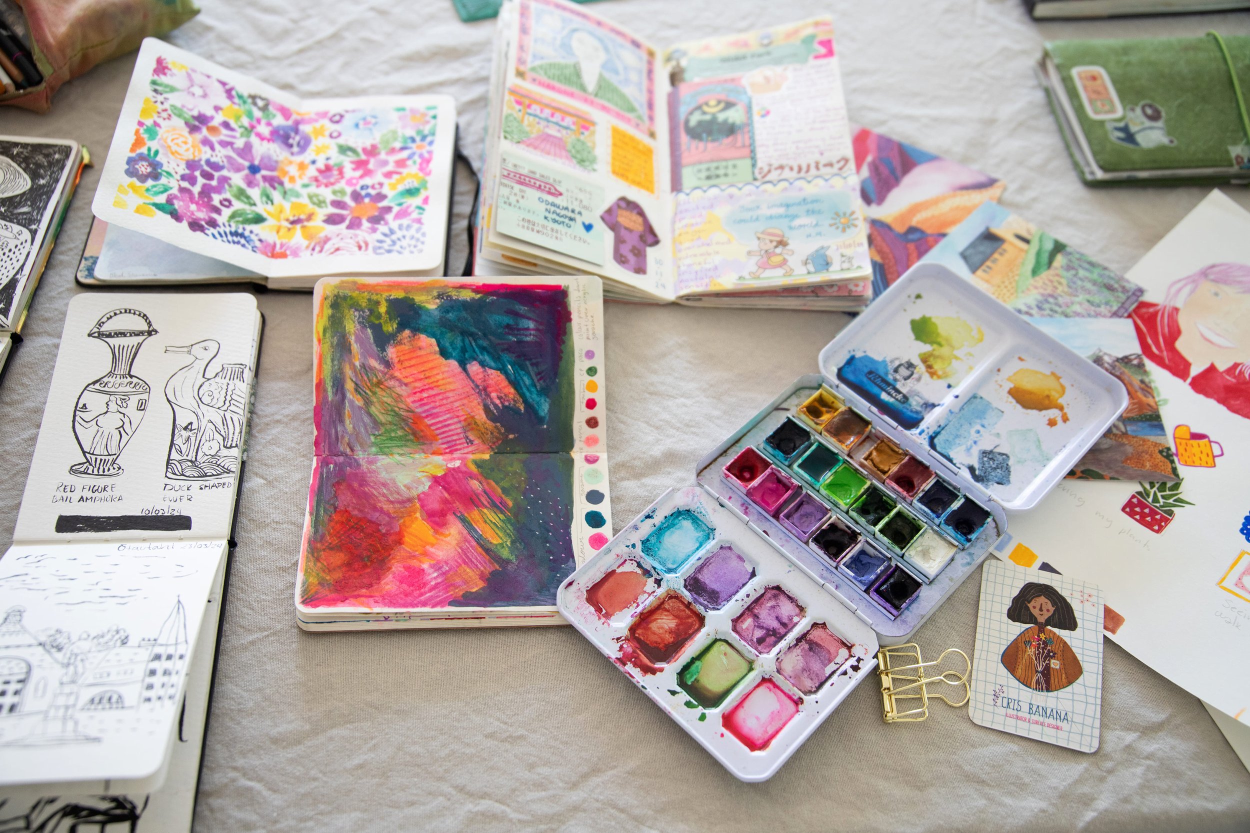 Open sketchbooks and watercolor paints on a table, displaying colorful artwork and illustrations.