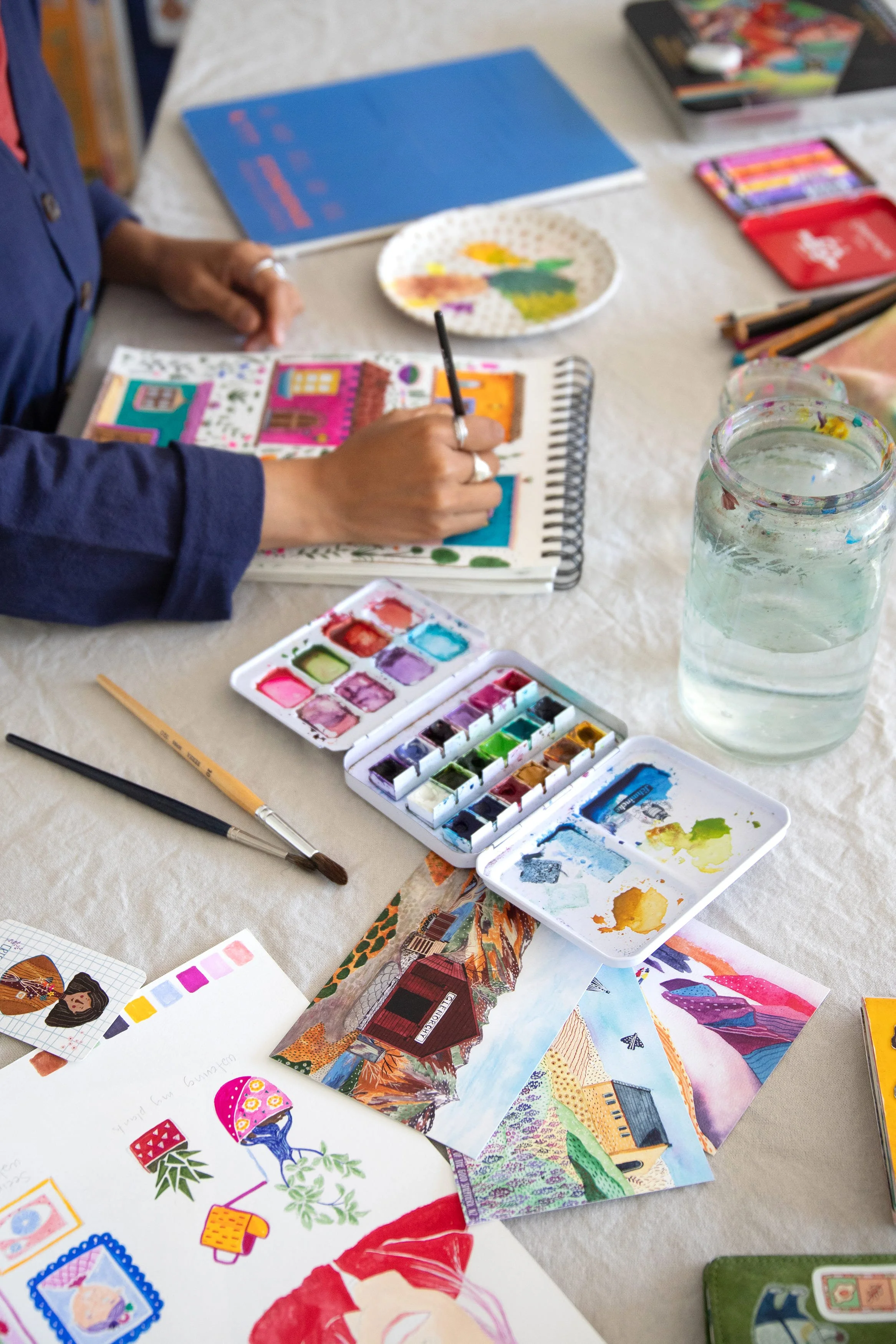 Person painting colorful illustrations and collages on paper with watercolor paints, surrounded by art supplies and water jars on a table.