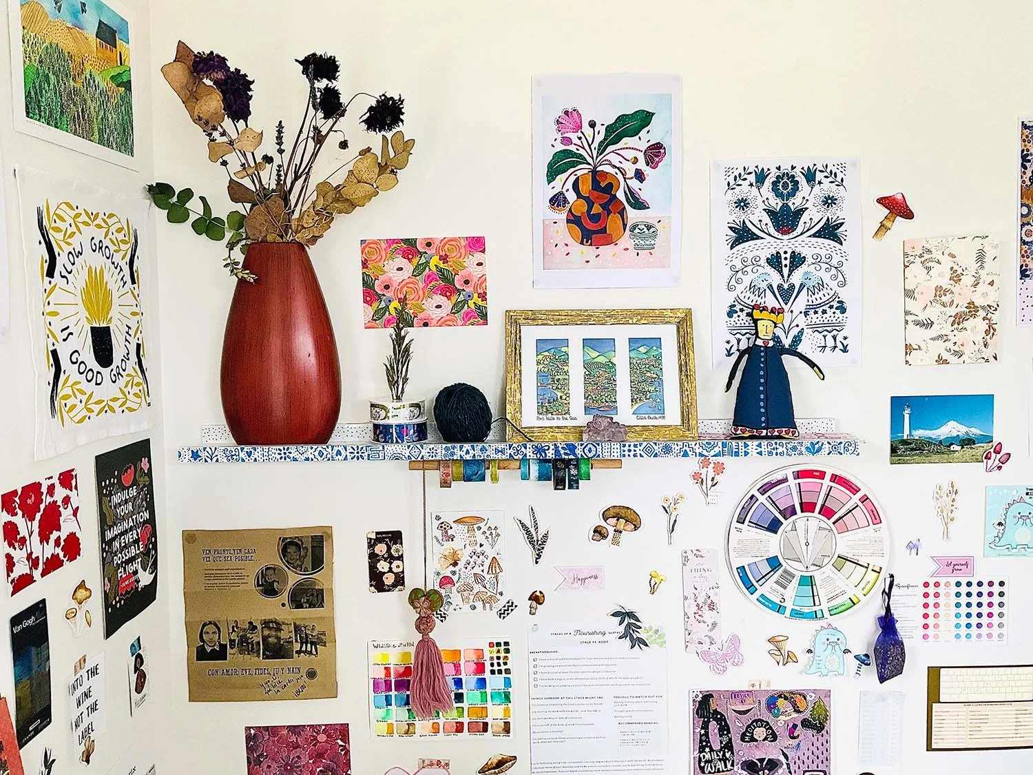 A wall decorated with various framed artwork, posters, and illustrations, including a large red vase with dried flowers on a shelf, a color palette wheel, and small decorative objects.