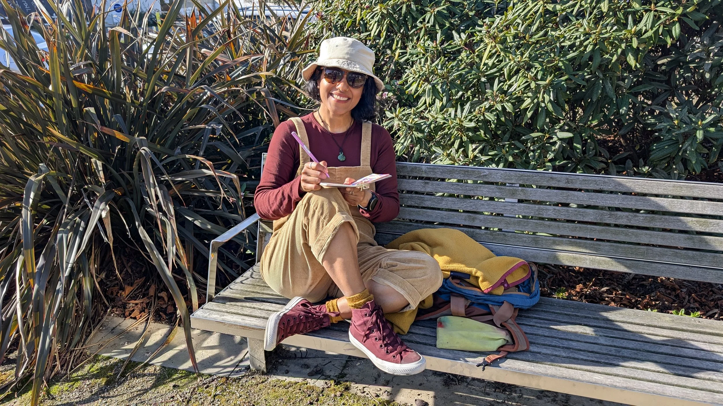 Woman sitting on a wooden bench outdoors, smiling, wearing sunglasses, a hat, and casual clothes, holding a notebook and pen, with backpacks and yellow blanket beside her, surrounded by plants.