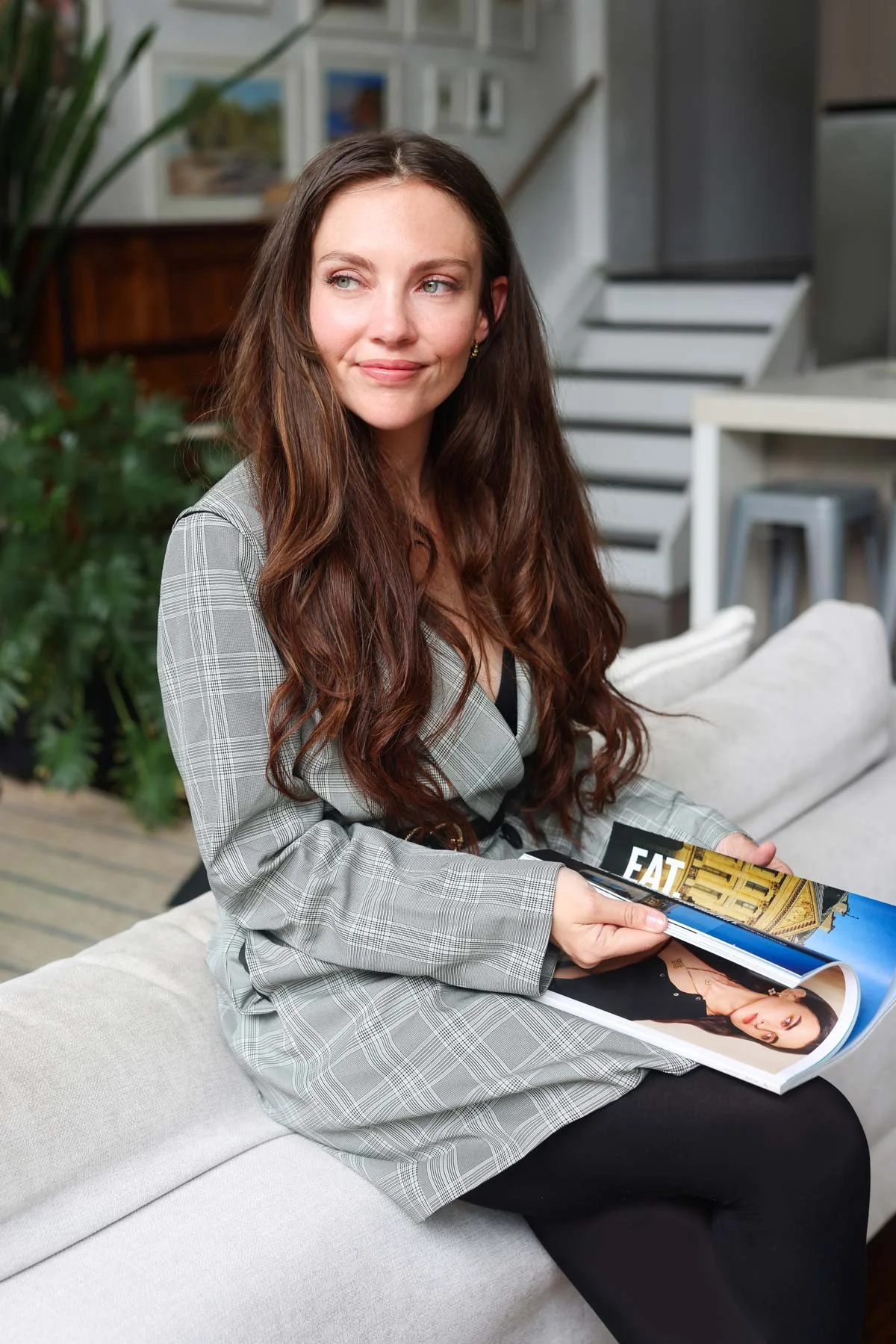 Emma Clark-Norris, progress expert, behavioural scientist; provisional psychologist; author; founder of Progress Magazine and The Progress Co. In checkered blazer sitting on a couch, holding a magazine, in a modern interior setting.