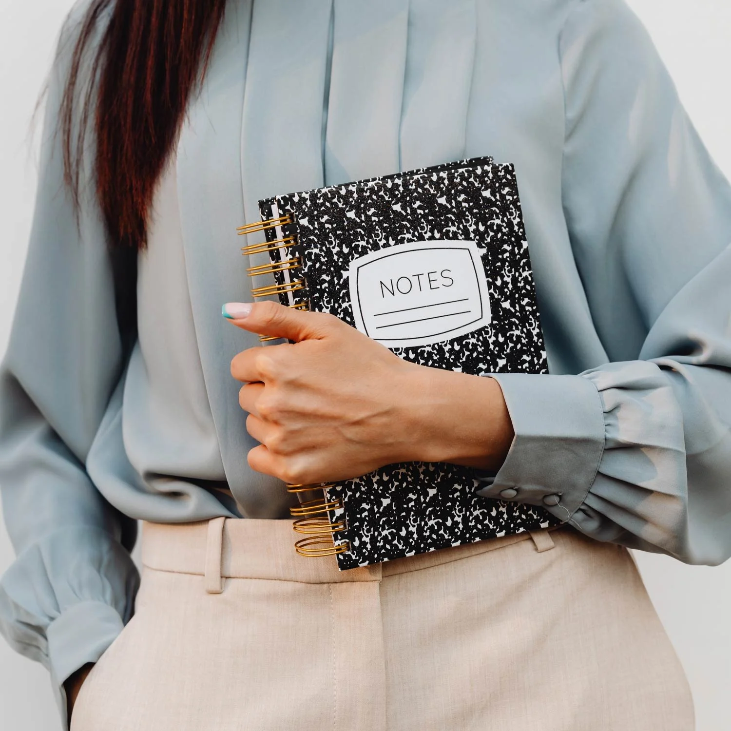 Person wearing a light blue blouse and beige pants holding a black and white spiral notebook labeled 'NOTES'.
