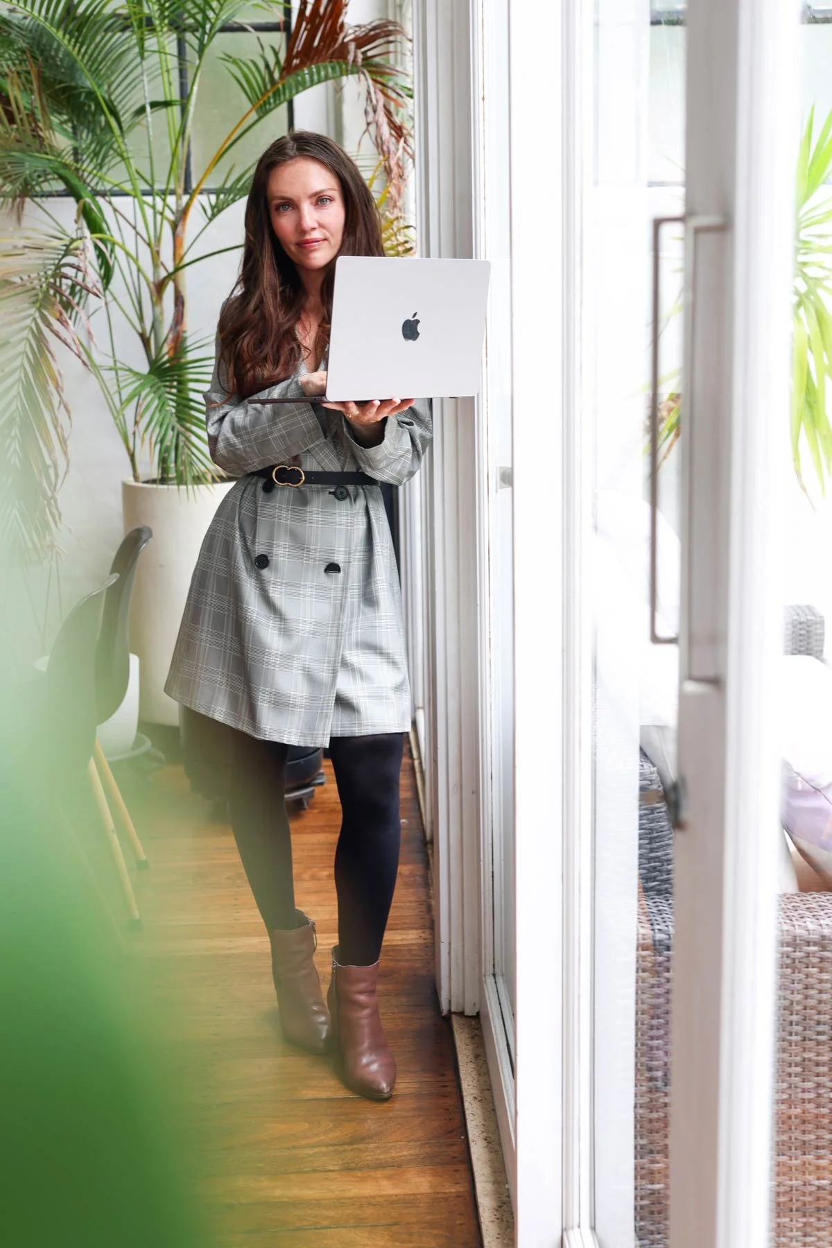 Emma Clark-Norris, progress expert, behavioural scientist; provisional psychologist; author; founder of Progress Magazine and, holding an open Apple MacBook laptop and standing indoors near a glass door with natural light, surrounded by green plants.
