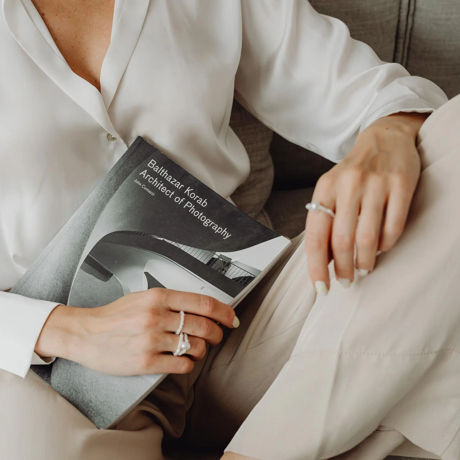 Close up detail shot of a person holding a book titled 'Balthazar Korab Architect or Photography,' wearing a white shirt, sitting on a gray couch.