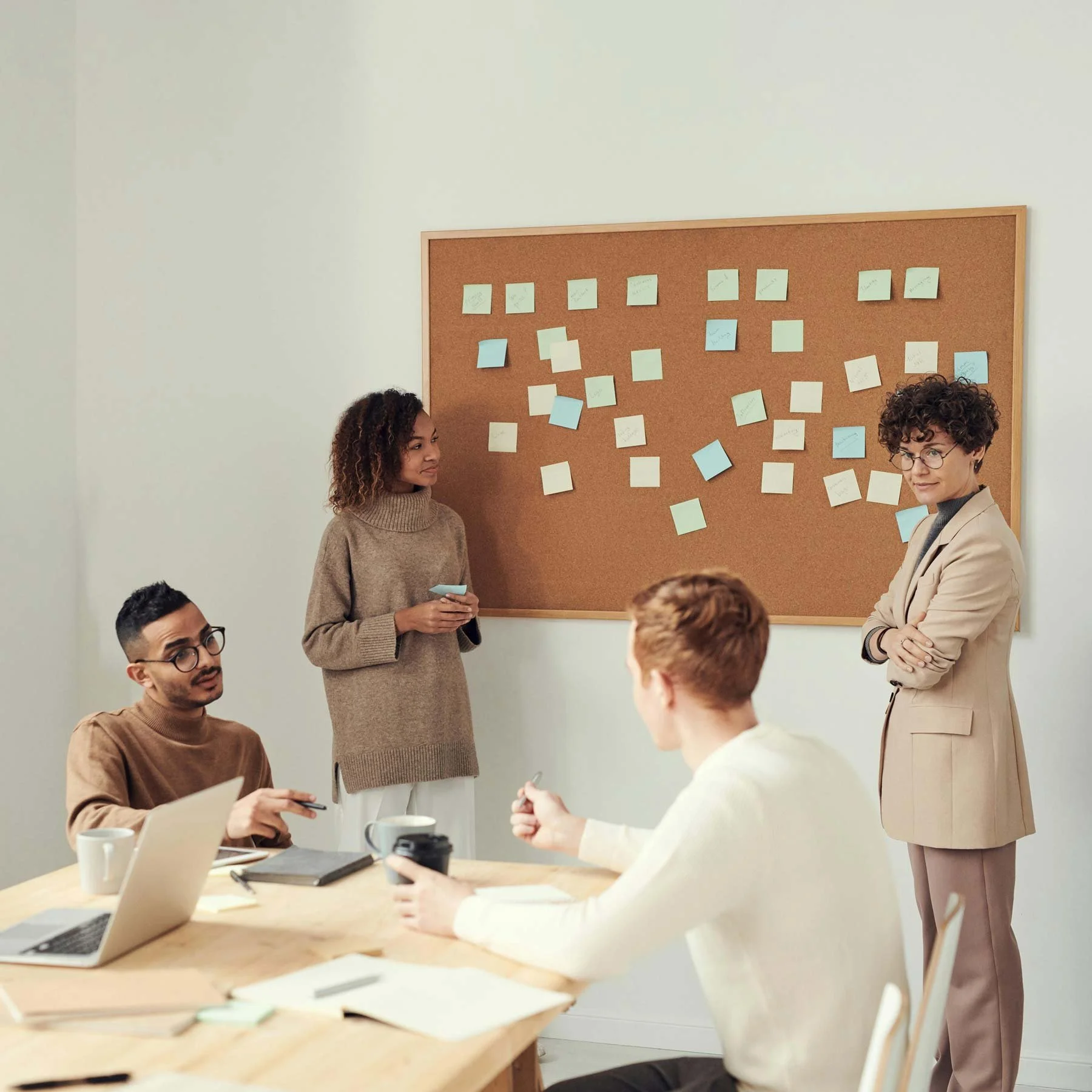 Four people in a meeting room discussing around a rectangular table with laptops, notebooks, and cups of coffee. A woman stands near a corkboard filled with colorful sticky notes. The room has white walls and modern decor.