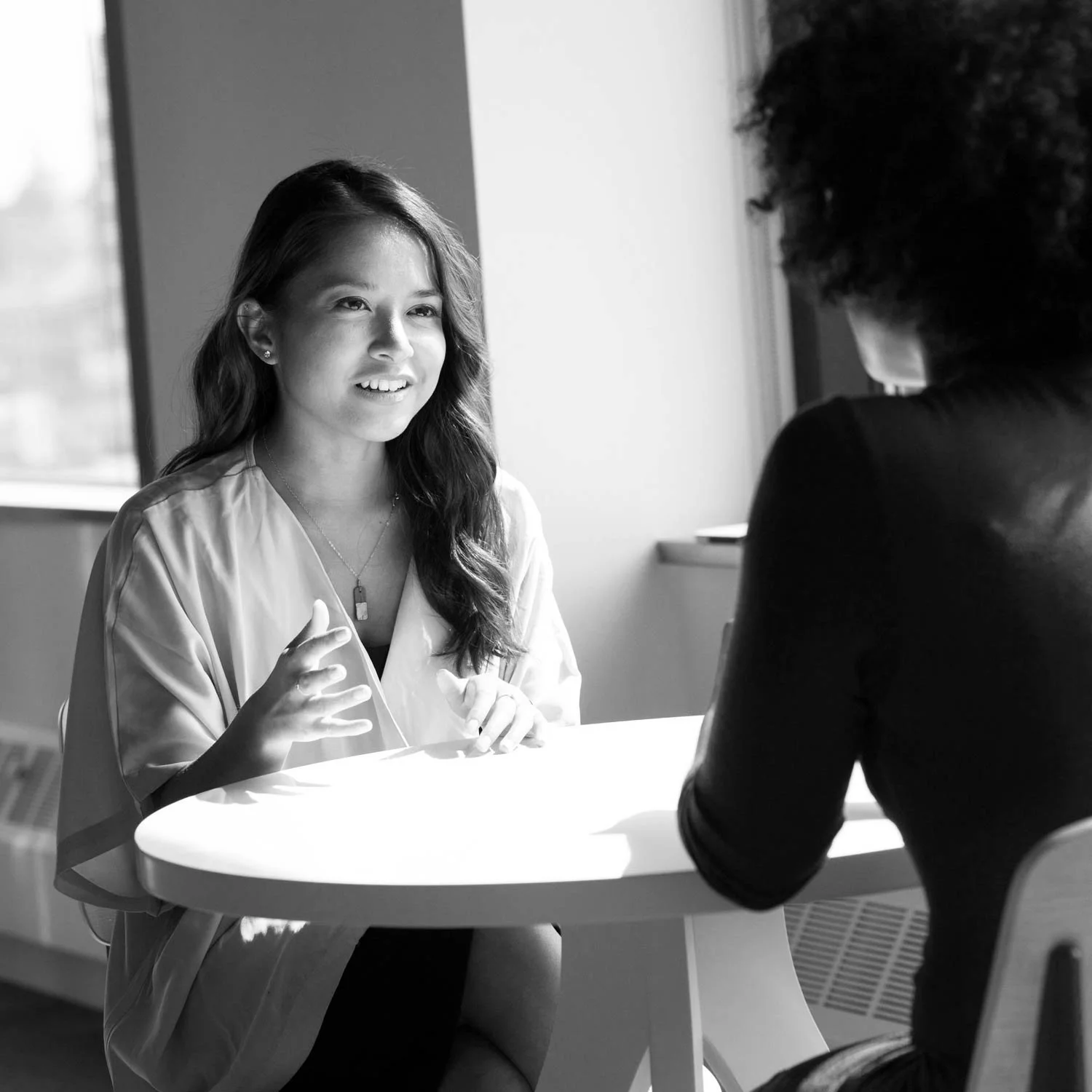 Two women having a conversation at a table in a bright room. One woman is smiling and gesturing, the other woman has curly hair and is listening.
