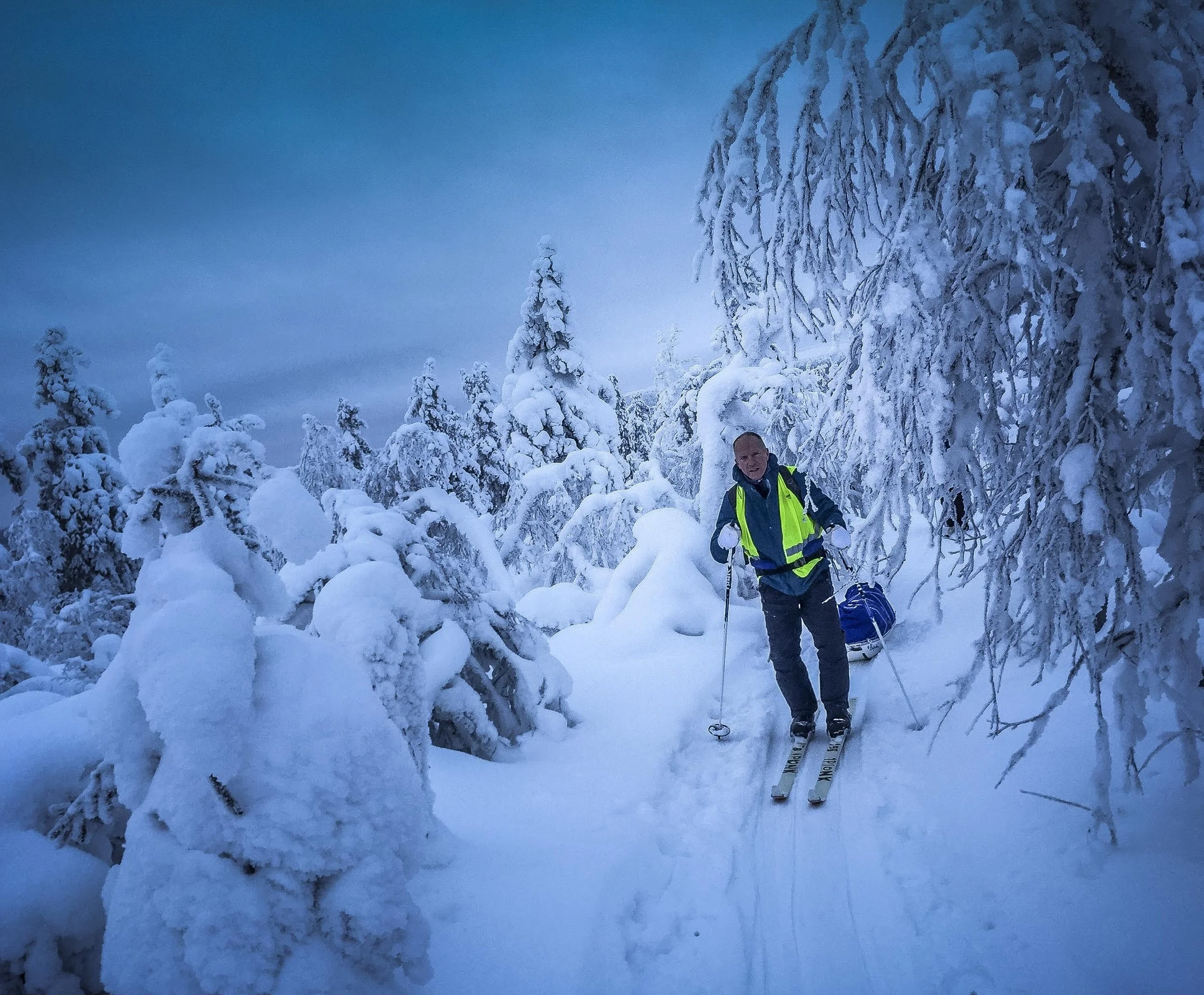 A person wearing a bright yellow safety vest and dark clothing is skiing on a snow-covered trail through a forest of snow-laden trees. The person is smiling and appears to be enjoying the winter scenery.