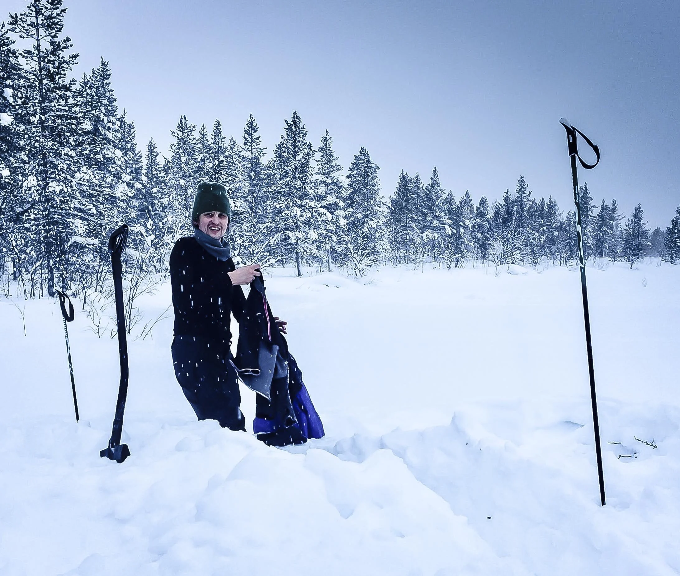 A person standing in deep snow in a winter landscape with snow-covered trees, smiling and holding a backpack, dressed in winter clothes.