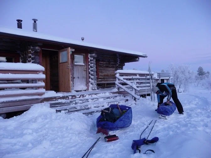 A person packing gear outside a snow-covered log cabin during winter, with snowshoes and a blue bag on the ground.