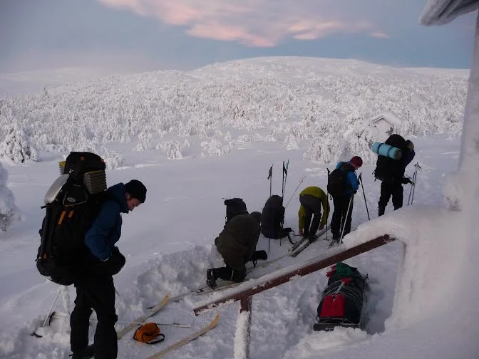 Group of people in winter clothing with ski equipment and backpacks trekking through snow near a snow-covered hill in a winter landscape.
