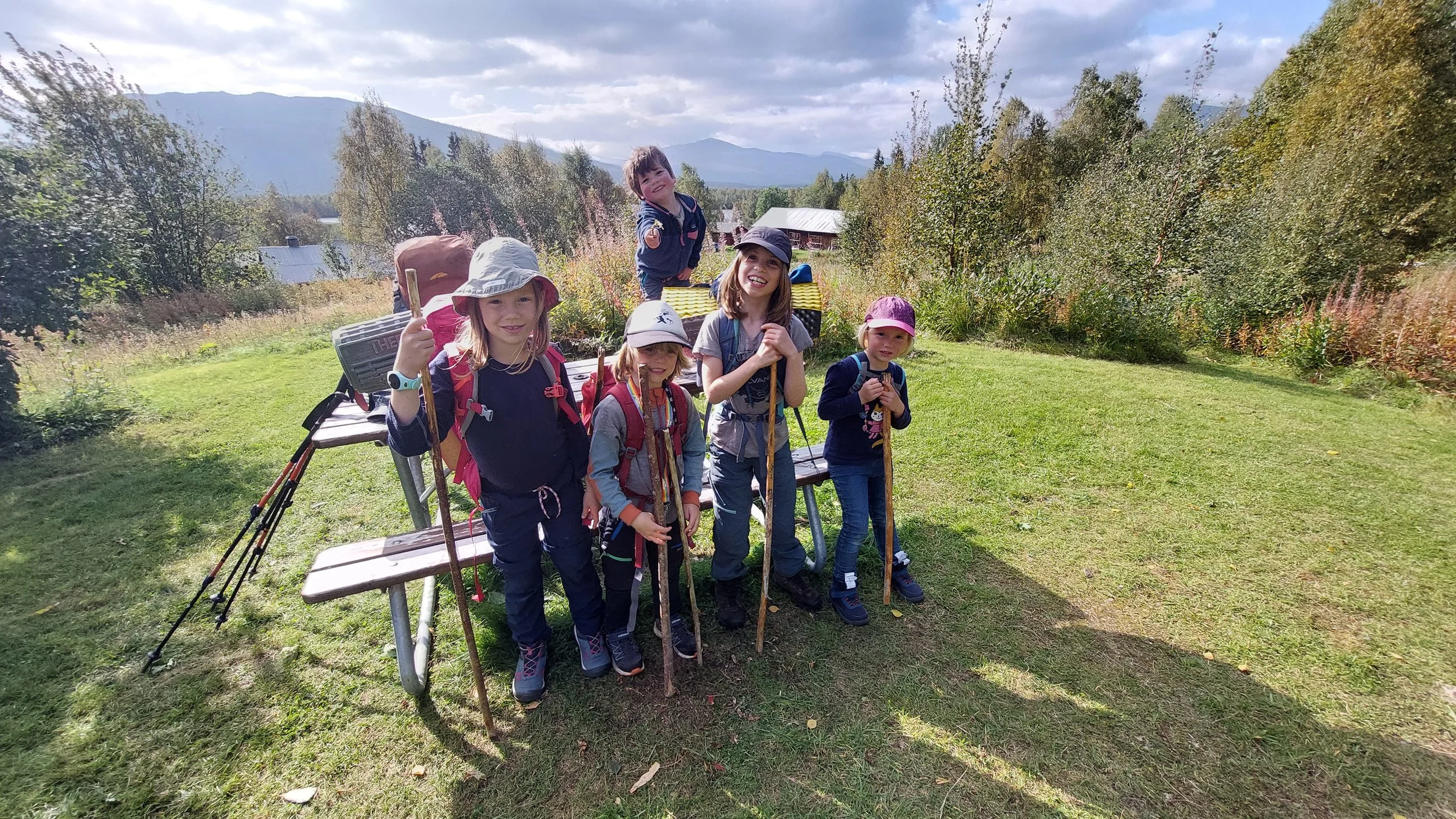 A group of six smiling children with backpacks and hiking sticks standing on a grassy area outdoors under a partly cloudy sky, with mountains and trees in the background.
