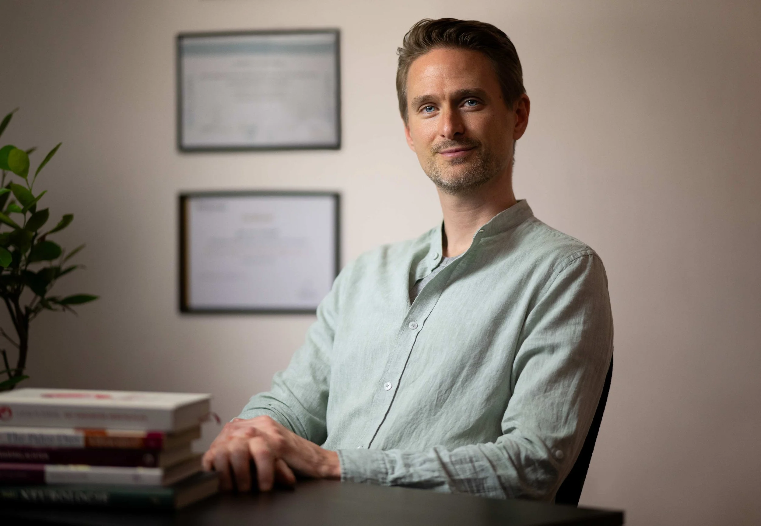 Jerome Verbeek in zijn praktijkruimte achter het bureau, groene blouse, met boeken en diploma’s op de achtergrond.
