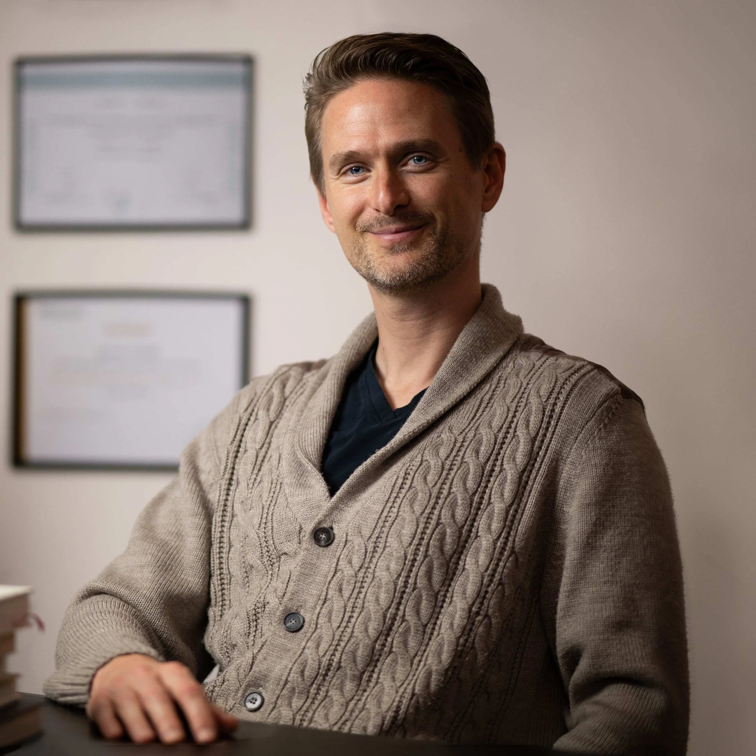 Jerome Verbeek aan zijn bureau in de praktijkruimte, bruin vest, met boeken en diploma’s op de achtergrond.