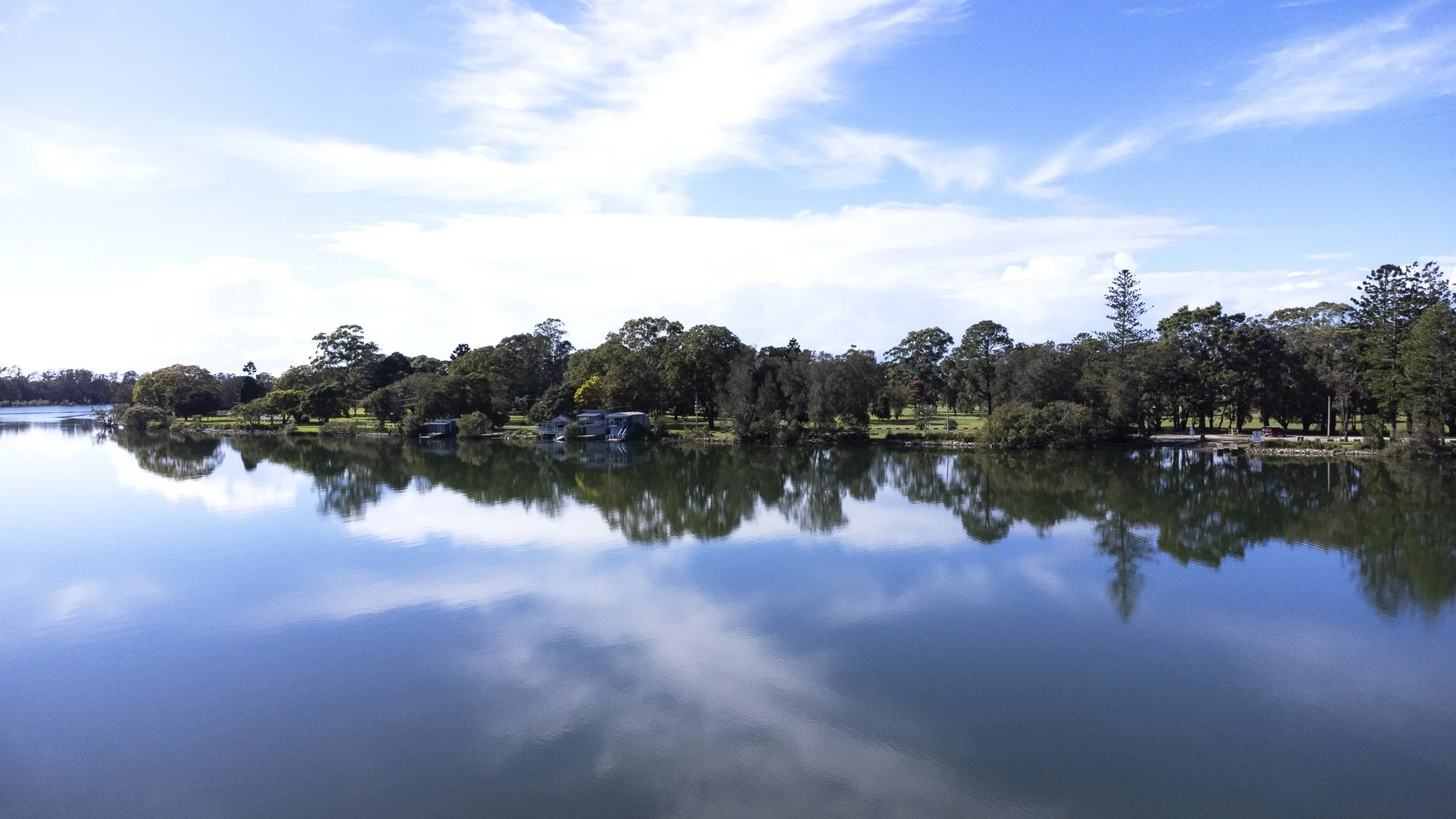 A peaceful lake scene with calm water reflecting a clear sky with some clouds, and a tree-lined shoreline with houses and trees.