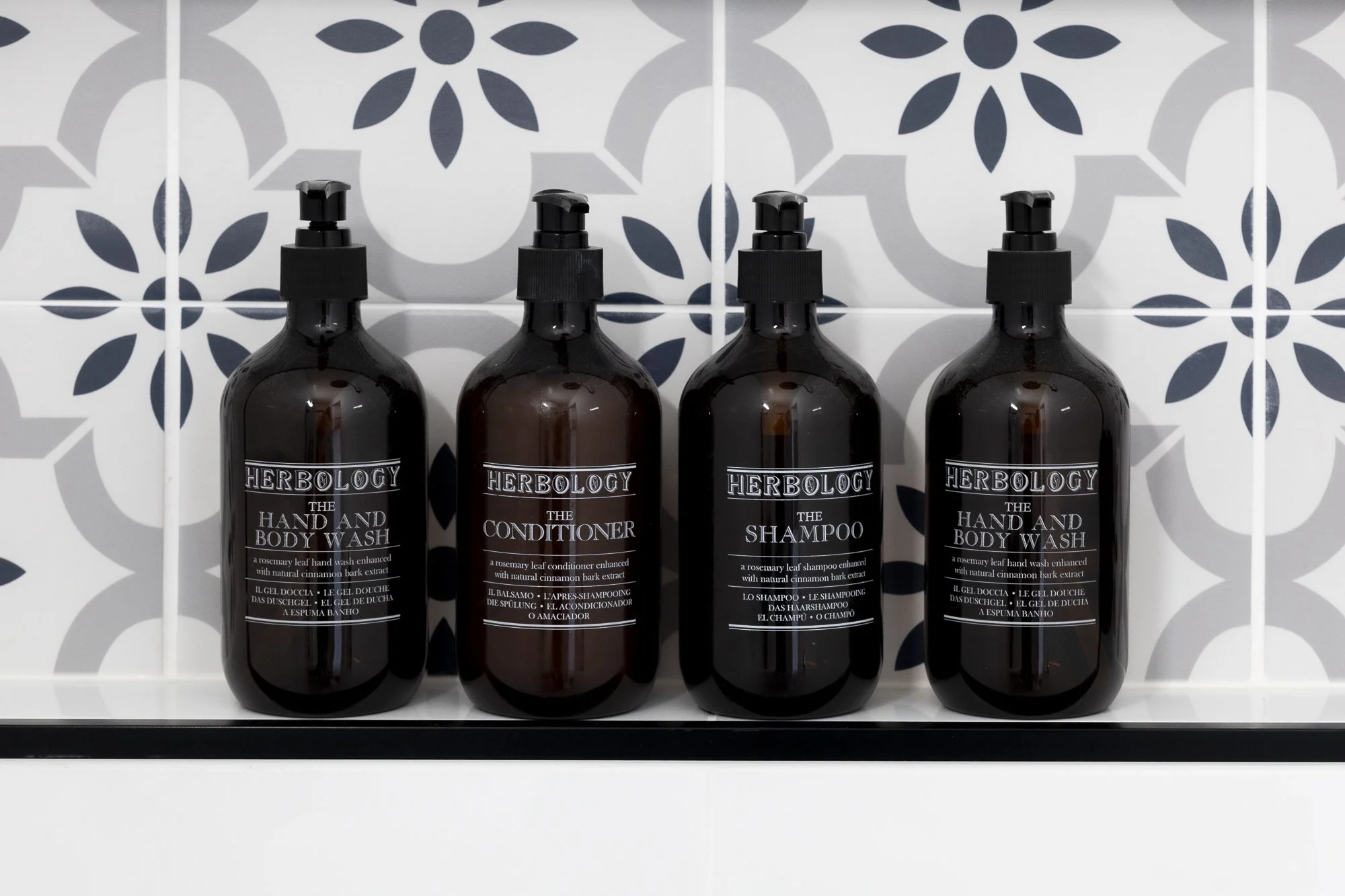 Four dark brown bottles of Herbolgy hair and body wash, conditioner, and shampoo with black pump dispensers, lined up on a black shelf against a tiled wall with a floral pattern.
