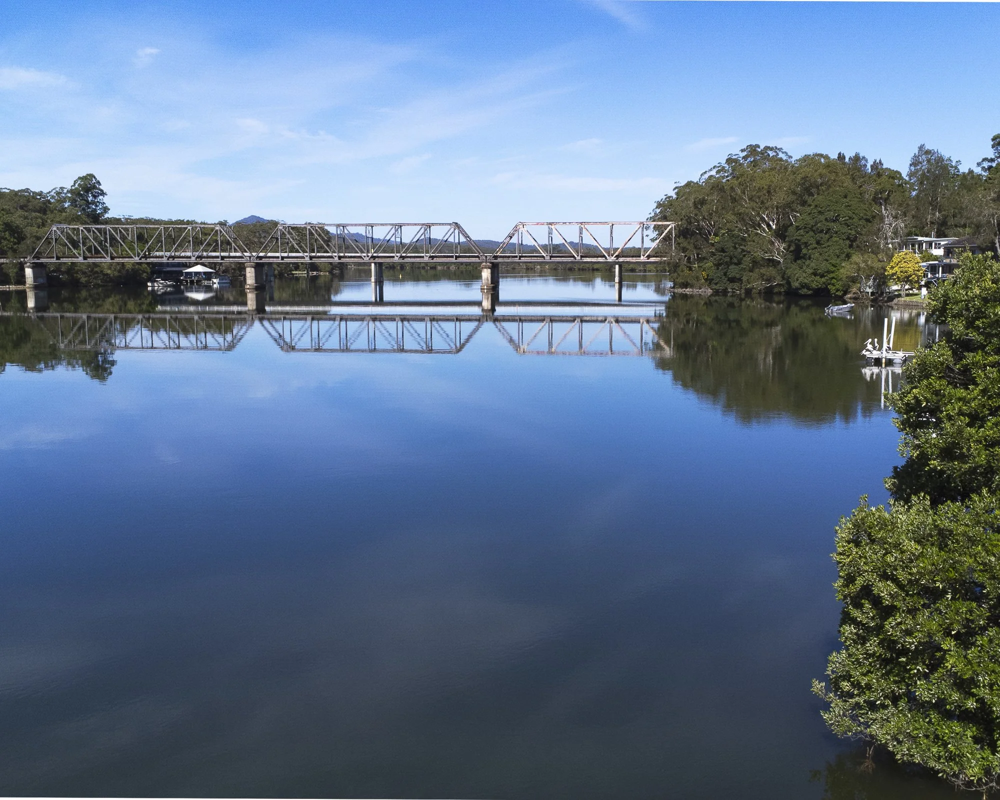 A calm river with a metal bridge crossing over it. The water reflects the bridge, sky, and surrounding trees. There are boats docked along the banks and lush green trees on both sides.