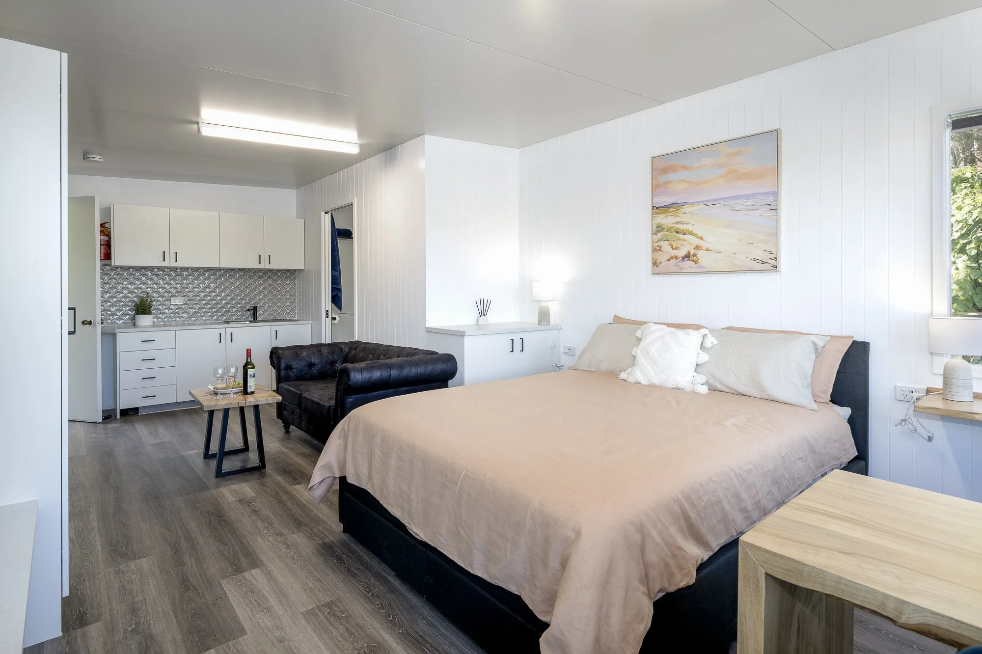 A bedroom with a large bed covered in beige bedding, two pillows, and a white decorative pillow. There is a painting of a beach scene hanging above the bed. To the right of the bed is a small nightstand with a lamp. On the left side, a black leather 