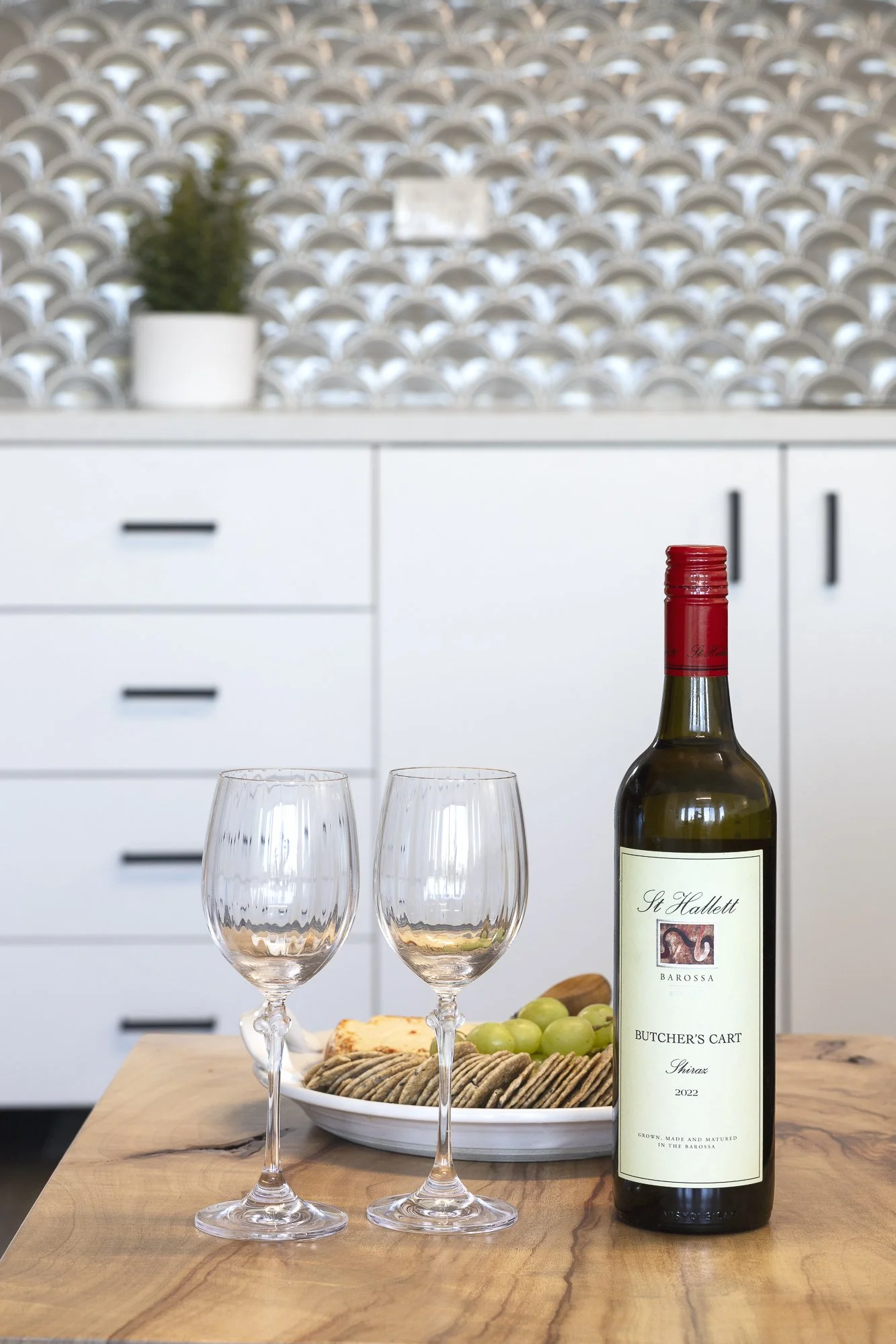 A bottle of red wine, two empty wine glasses, a bowl of cheese, grapes, and crackers on a wooden table in a modern kitchen.