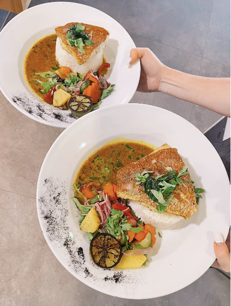 Two plates of fish and rice served with mixed vegetables and lemon slices, garnished with herbs, on a table.