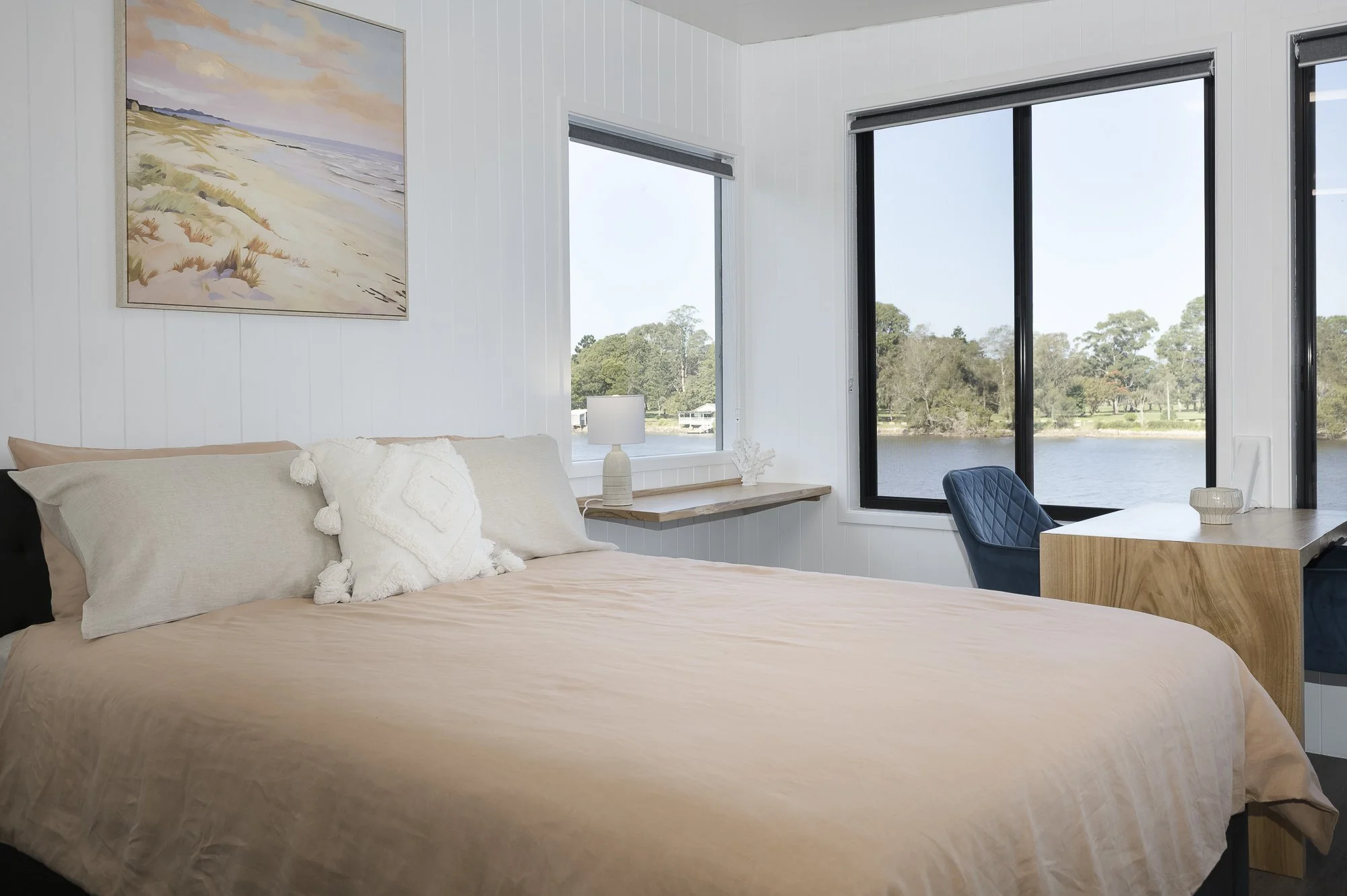 A bedroom with white shiplap walls and large windows overlooking a lake and trees. It has a large bed with beige bedding, decorative pillows, a wooden desk with a blue chair, and a nightstand with a lamp. A landscape painting hangs above the bed.
