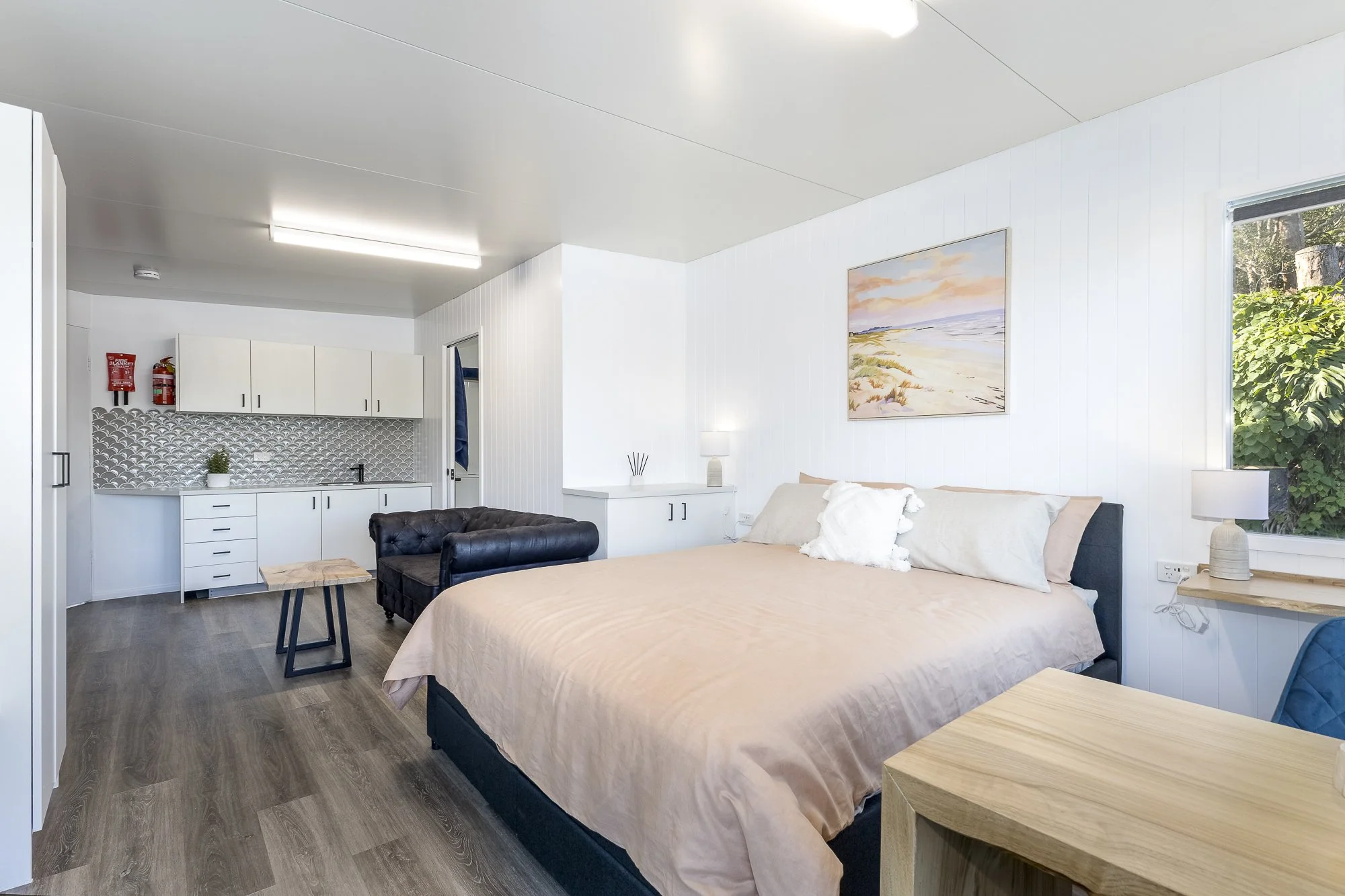Clean and modern bedroom with a large bed with beige bedding, a nightstand with lamp, a white cabinet, and a window with greenery outside. There is also a small kitchenette with white cabinets, a patterned backsplash, and a small table.