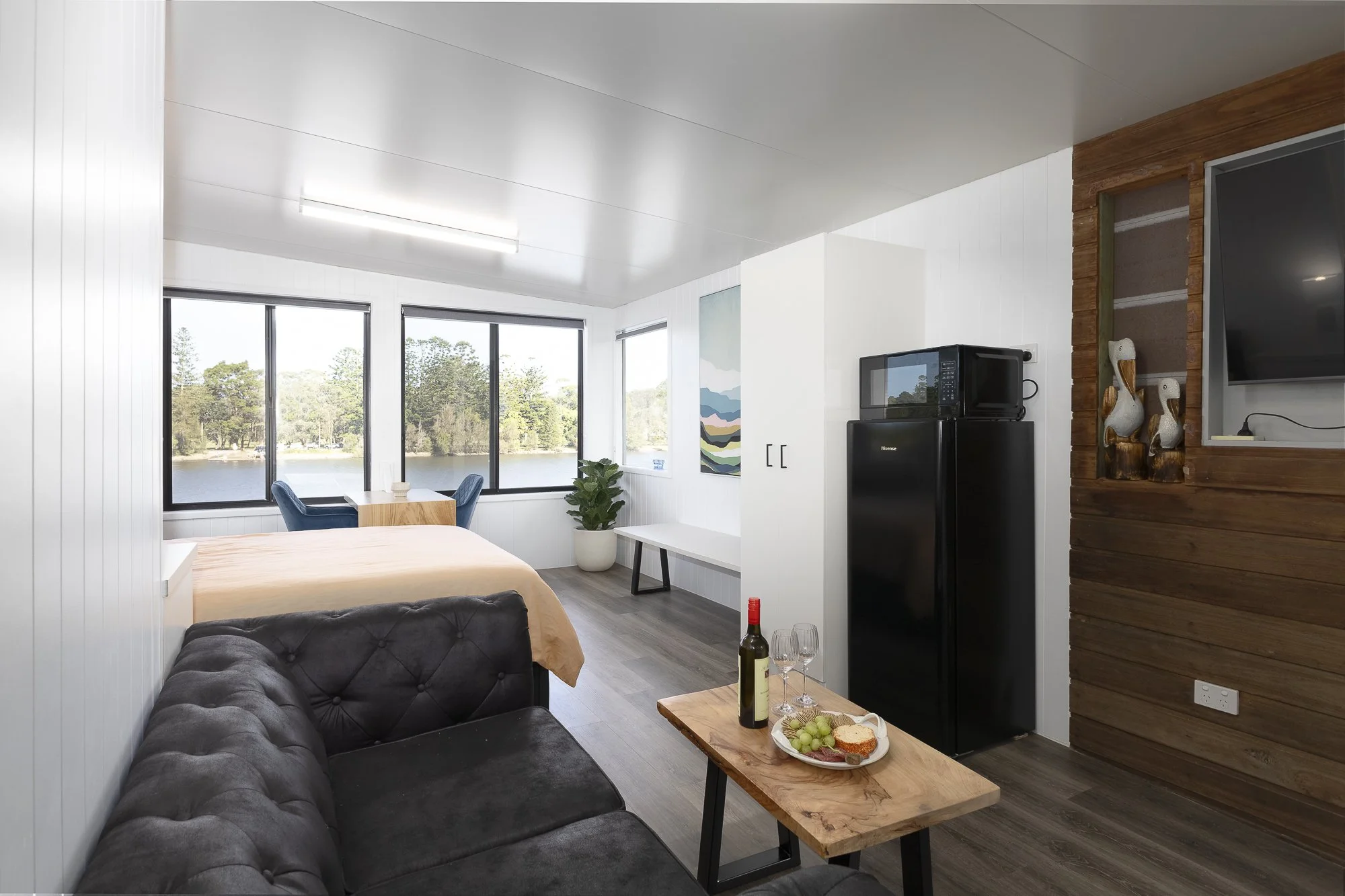 Interior of a modern studio apartment with a view of a lake through large windows, including a bed, a black sofa, a wooden coffee table with a bottle of wine and snacks, a white cabinet, a small table with a plant, and kitchen appliances.