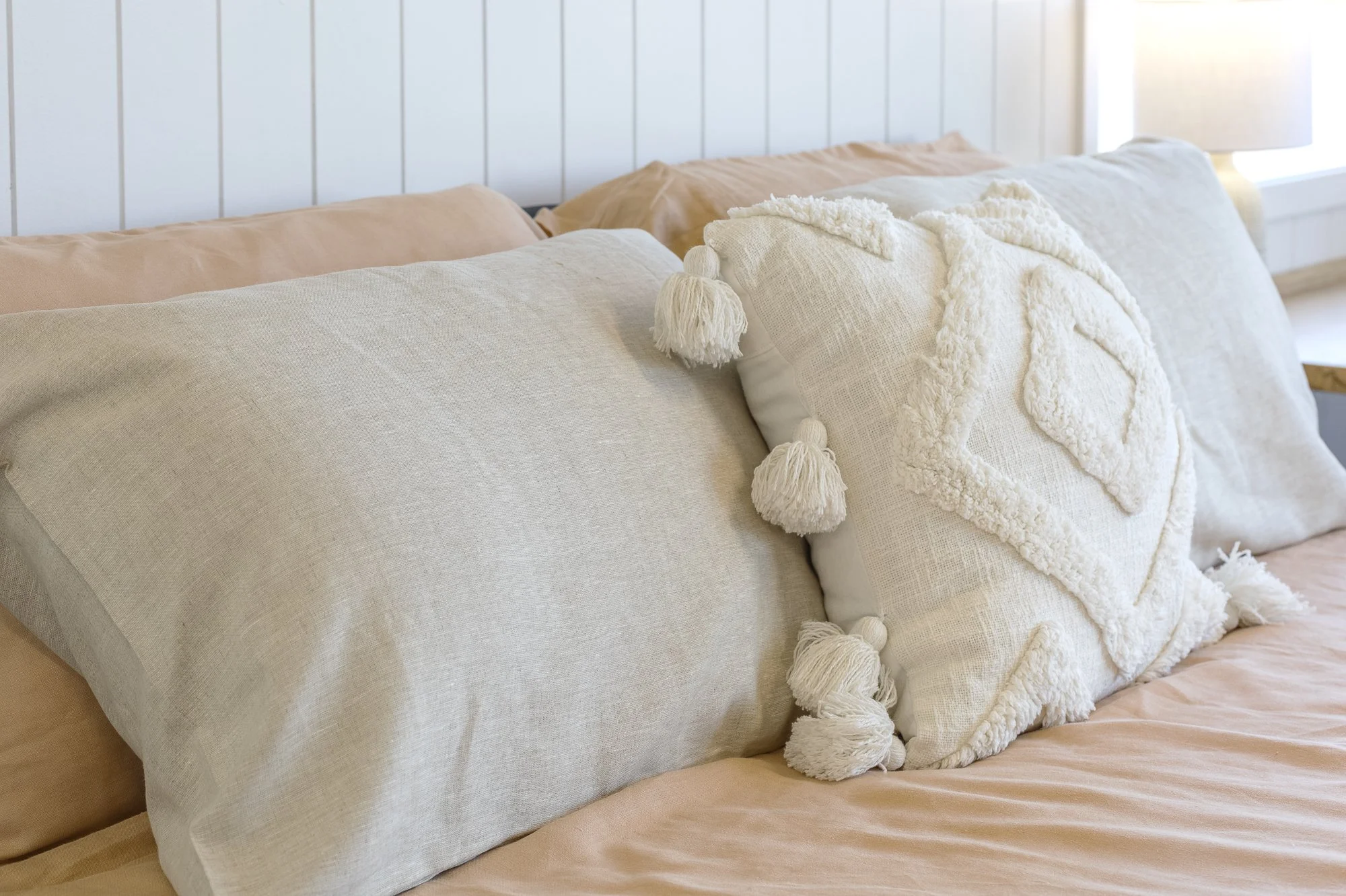 A neatly made bed with beige and white pillows, including a decorative white pillow with tassels and textured embroidery, against a white paneled wall with sunlight coming through a nearby window.