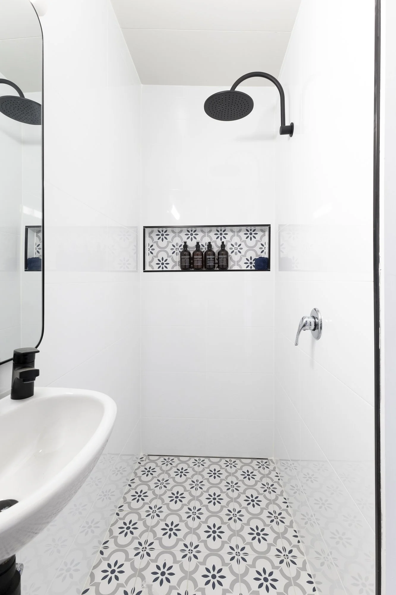 A minimalist shower area with a black overhead rain showerhead, white tiled walls, a built-in tiled niche holding brown bottles, and a decorative patterned tile floor.