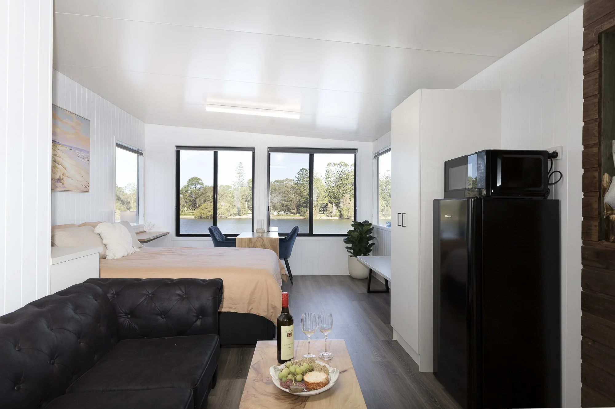 Interior of a small apartment with a bed, black sofa, wooden coffee table, wine bottle and glasses, kitchenette with microwave and refrigerator, and large windows overlooking a lake.
