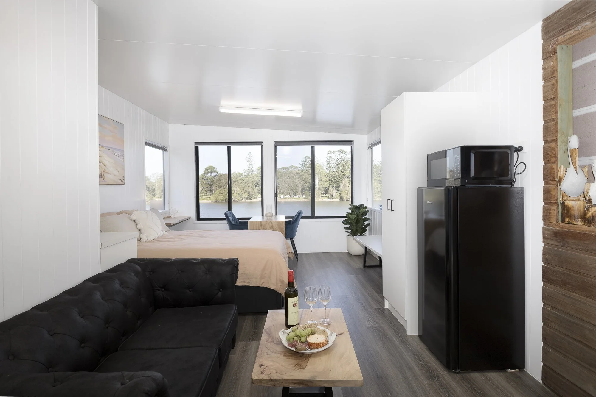 Interior view of a small modern apartment with a bed, a dining table with chairs, a black couch with a bottle of wine and glasses, a microwave, a mini fridge, a plant, and large windows overlooking a lake.