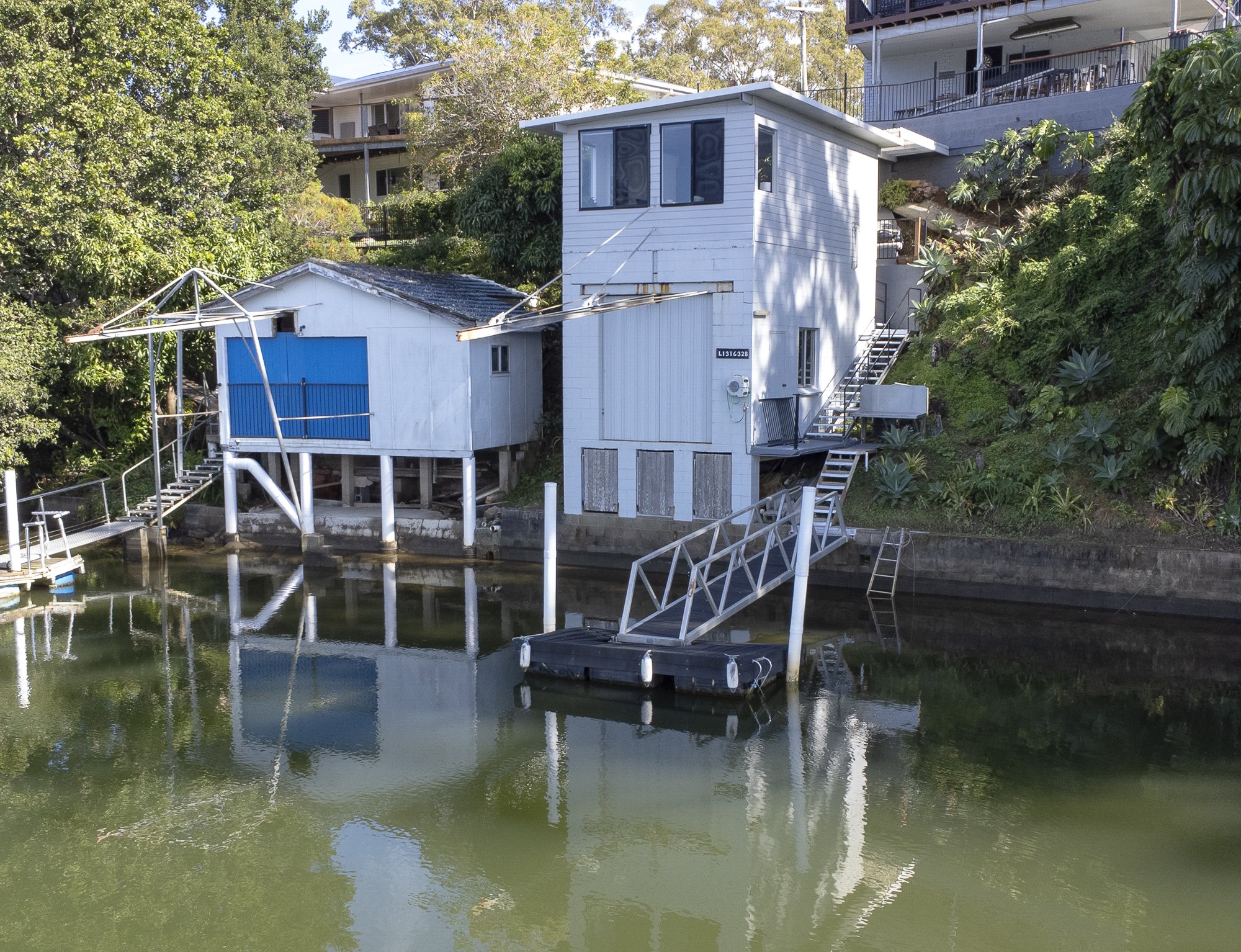 Two houses on stilts by a river, with boat lifts and staircases leading to the water, surrounded by greenery.