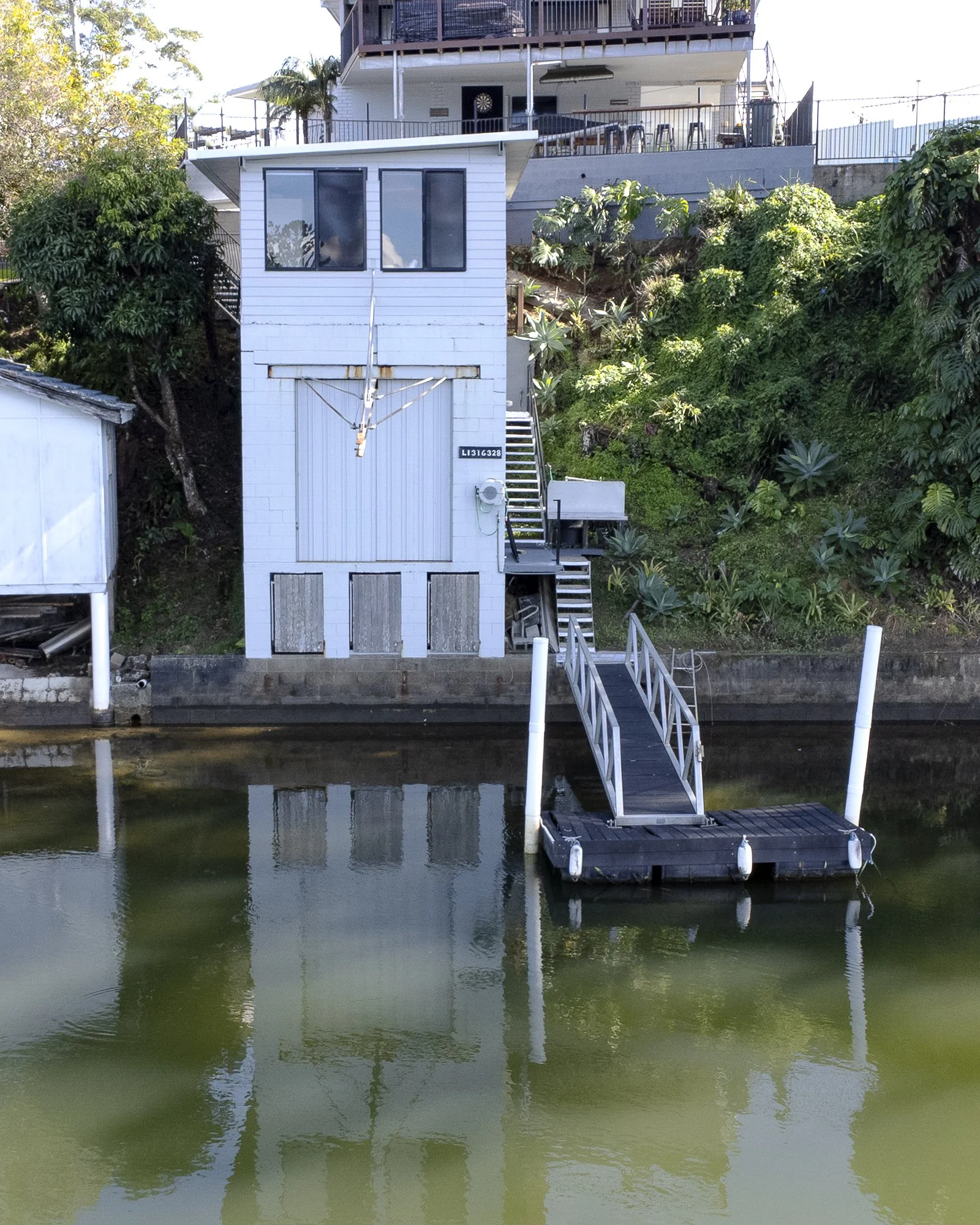A white building with a garage door and stairs leading to the top, situated on a waterfront with a dock extending over the water, and lush green vegetation on the hillside behind.