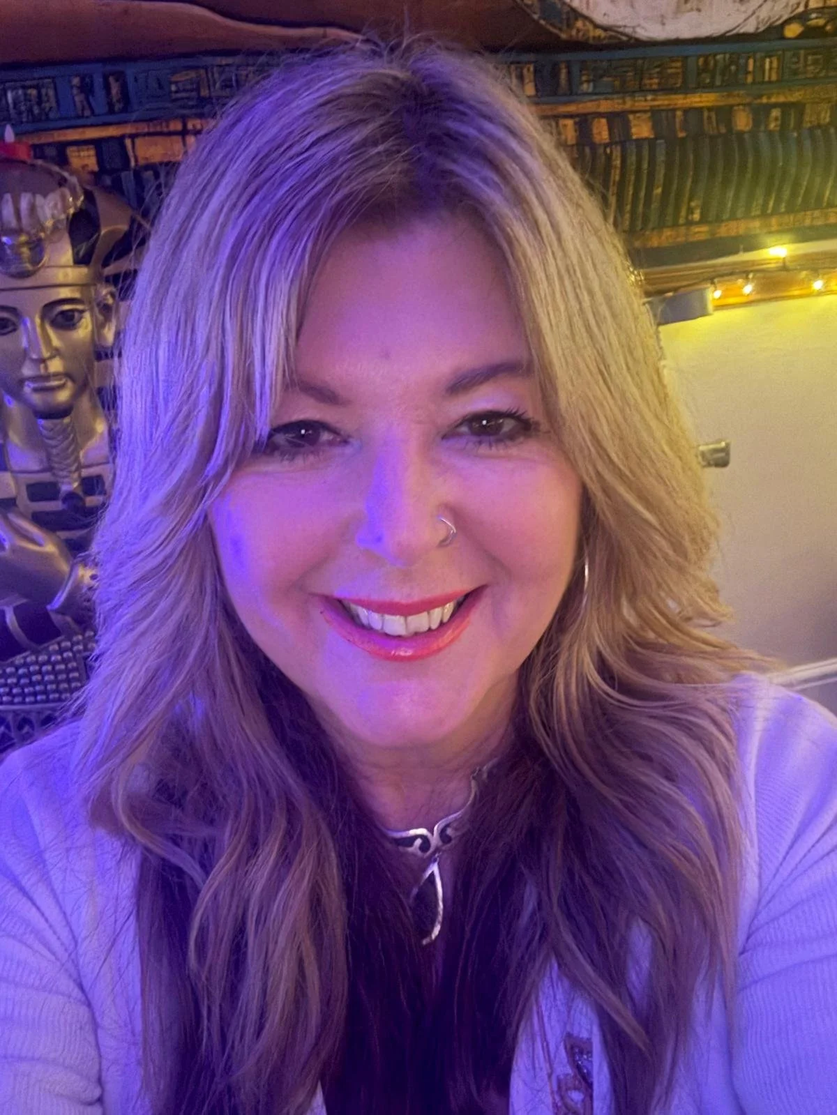 A woman with long wavy blonde hair smiling, indoor setting with purple lighting, some metallic decorations and a decorative object in the background.