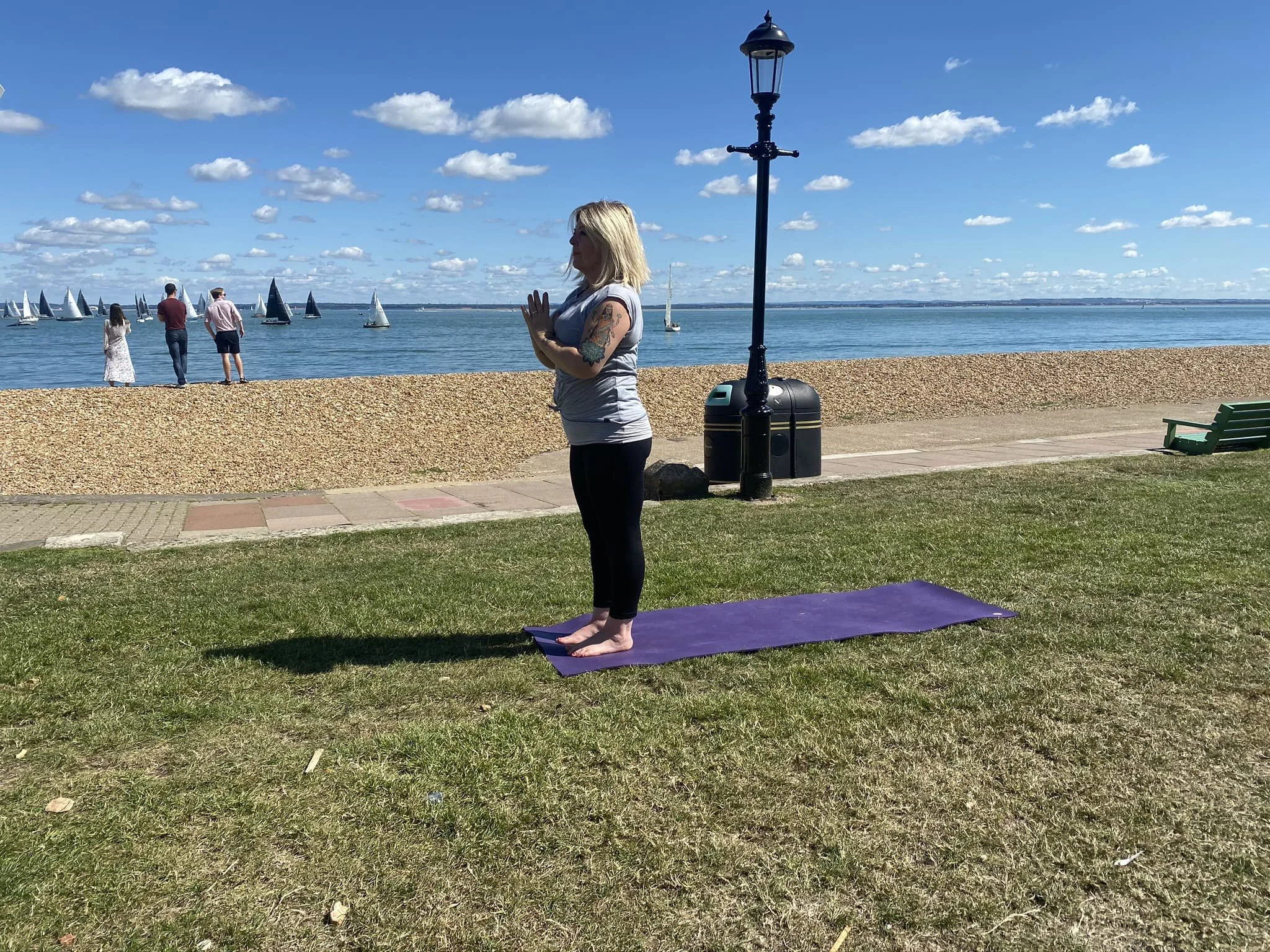 A woman practicing yoga outdoors on a purple mat near the water, with sailboats, a lamppost, and a bench in the background, on a sunny day with clouds in the sky.