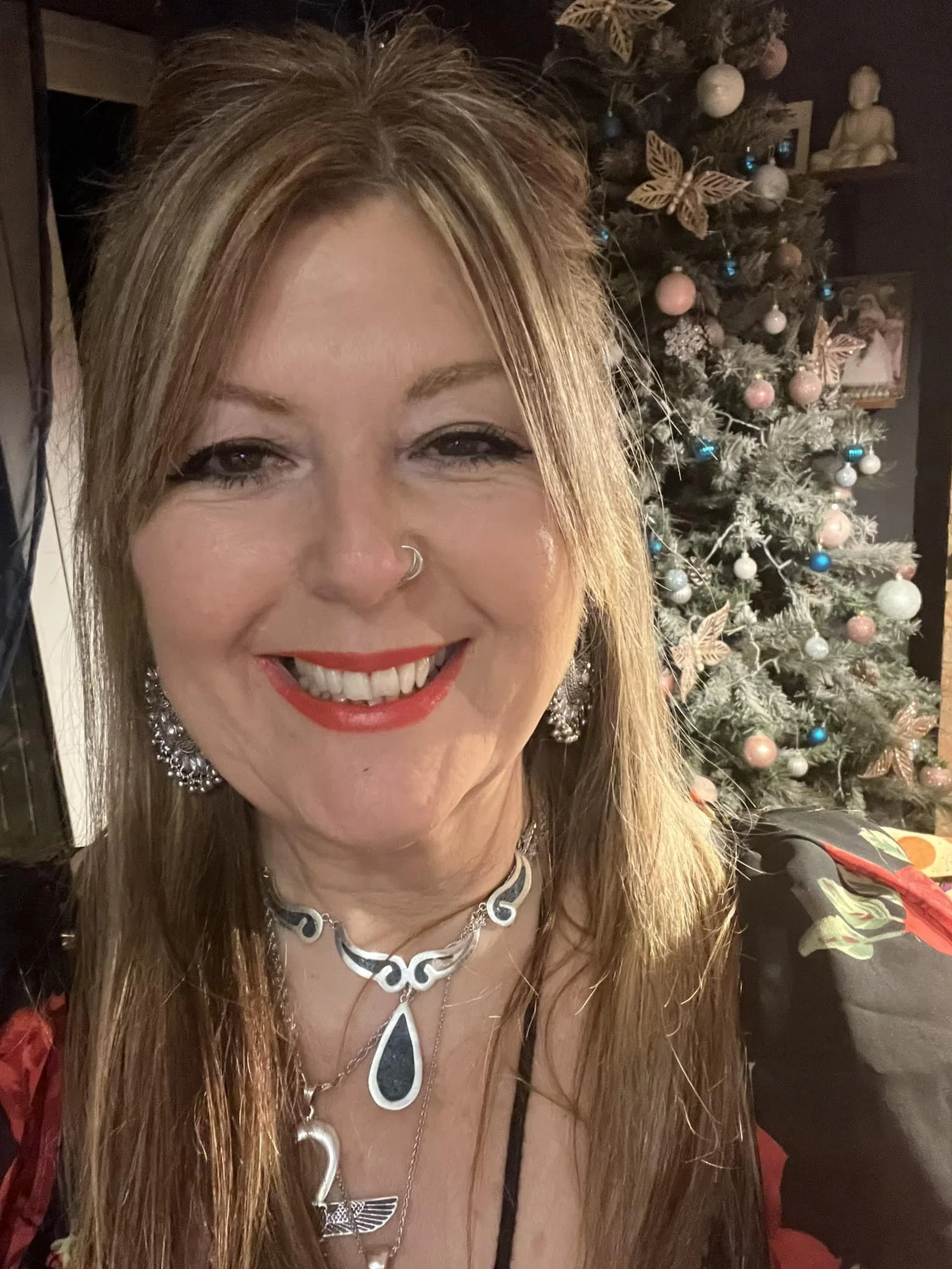 A woman smiling in front of a decorated Christmas tree, wearing jewelry and a floral dress.