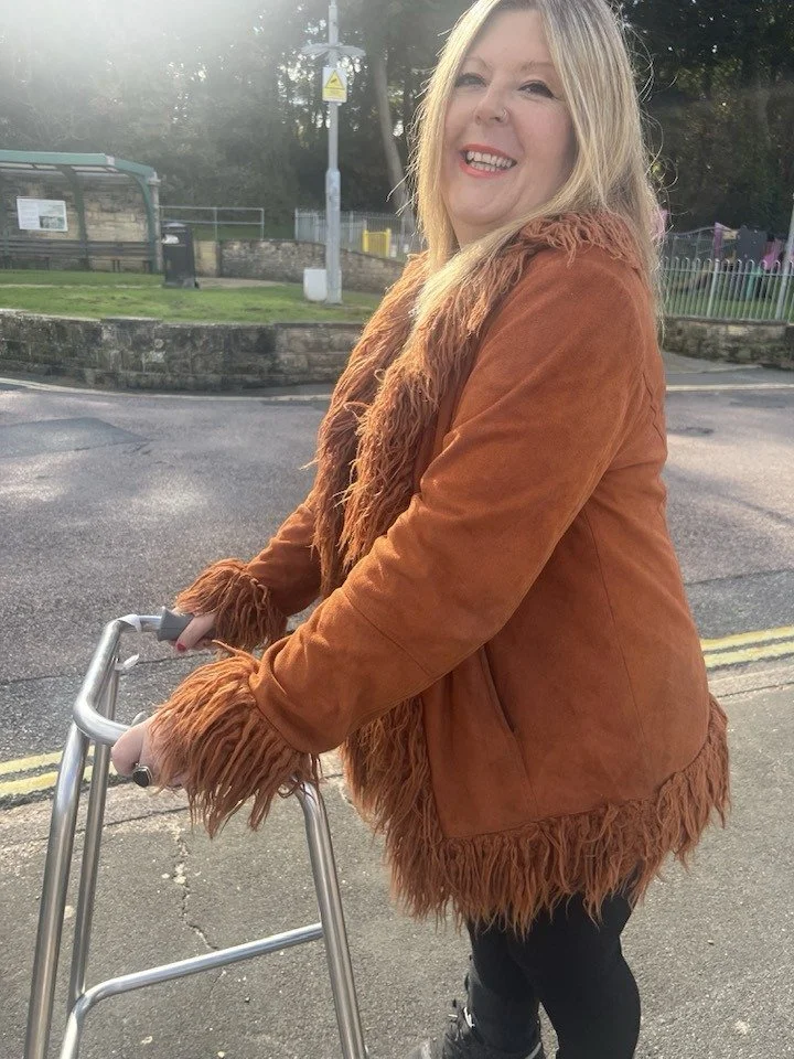 A woman with blonde hair smiling while standing outdoors, dressed in a brown suede jacket with fringes.