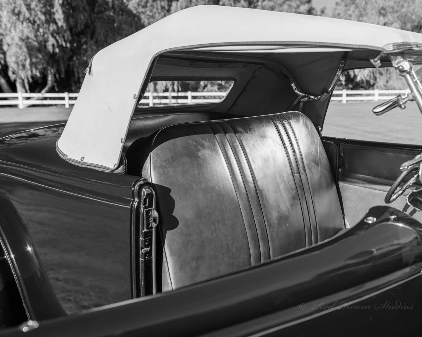 Black and white photo of vintage 1935 Ford Roadster car interior showing leather seats and open roof with a view of a fence and trees outside.