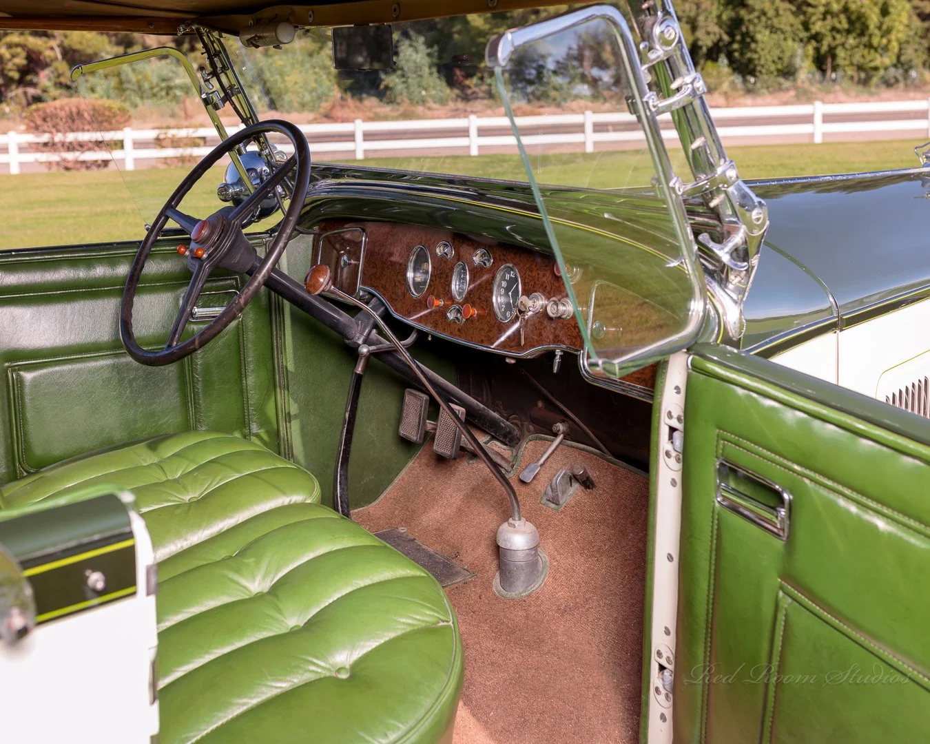 Interior of a vintage 1932 Packard with a green leather bench seat, wooden dashboard, and various gauges, with a large steering wheel and a windshield with chrome supports. Classic car experiences by Gabby Lane Classics.