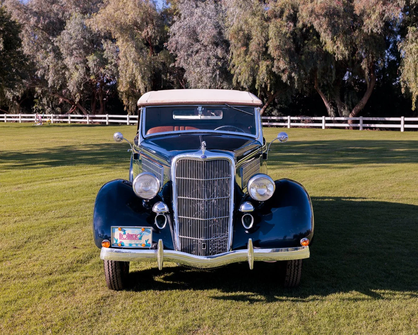 Front view of a black vintage 1935 Ford Roadster car parked on a grassy field with trees and a white fence in the background. Classic car experiences by Gabby Lane Classics.
