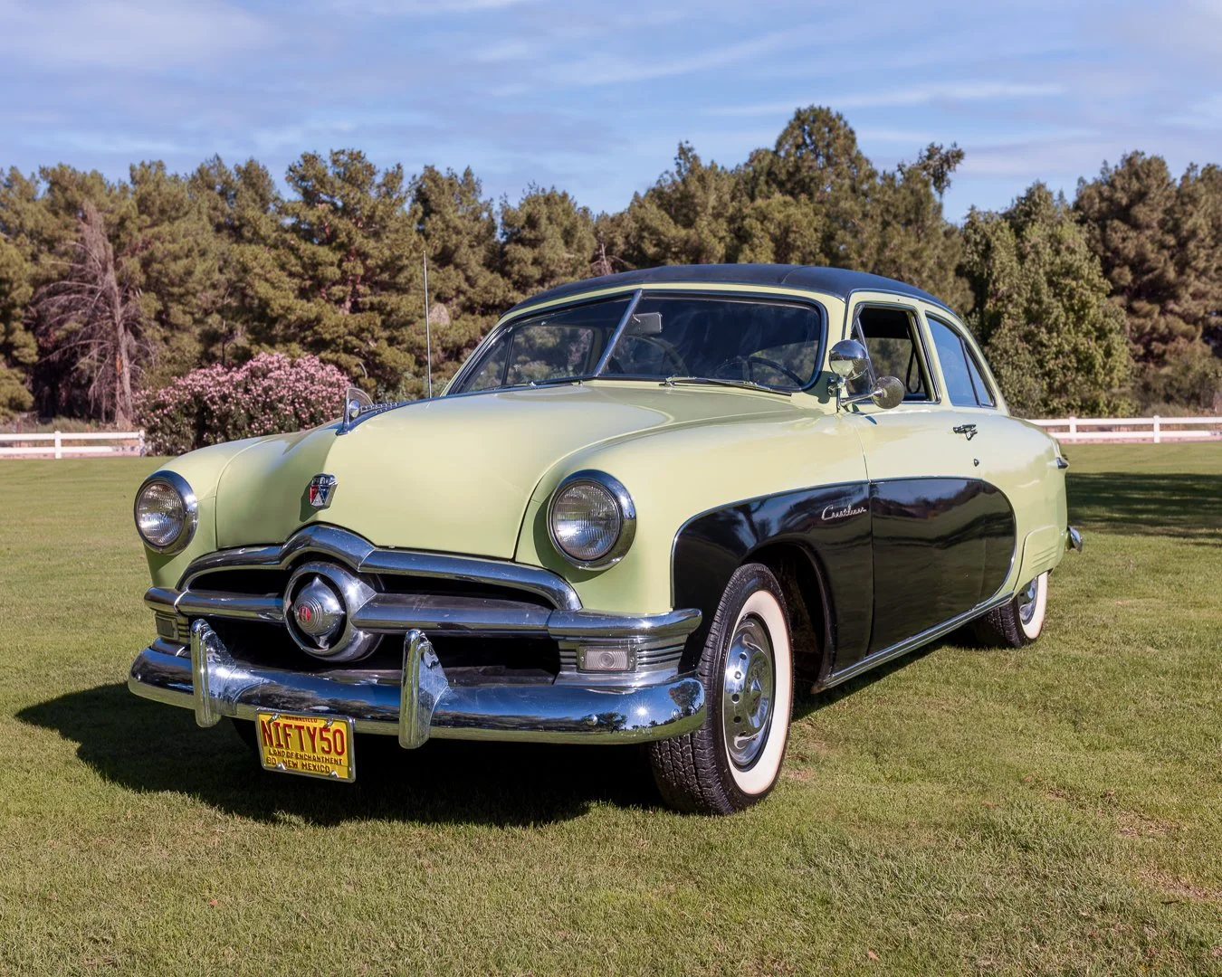 A vintage green and black 1950 Ford Crestliner parked on a grassy field with trees and a fence in the background. Classic car experiences by Gabby Lane Classics.